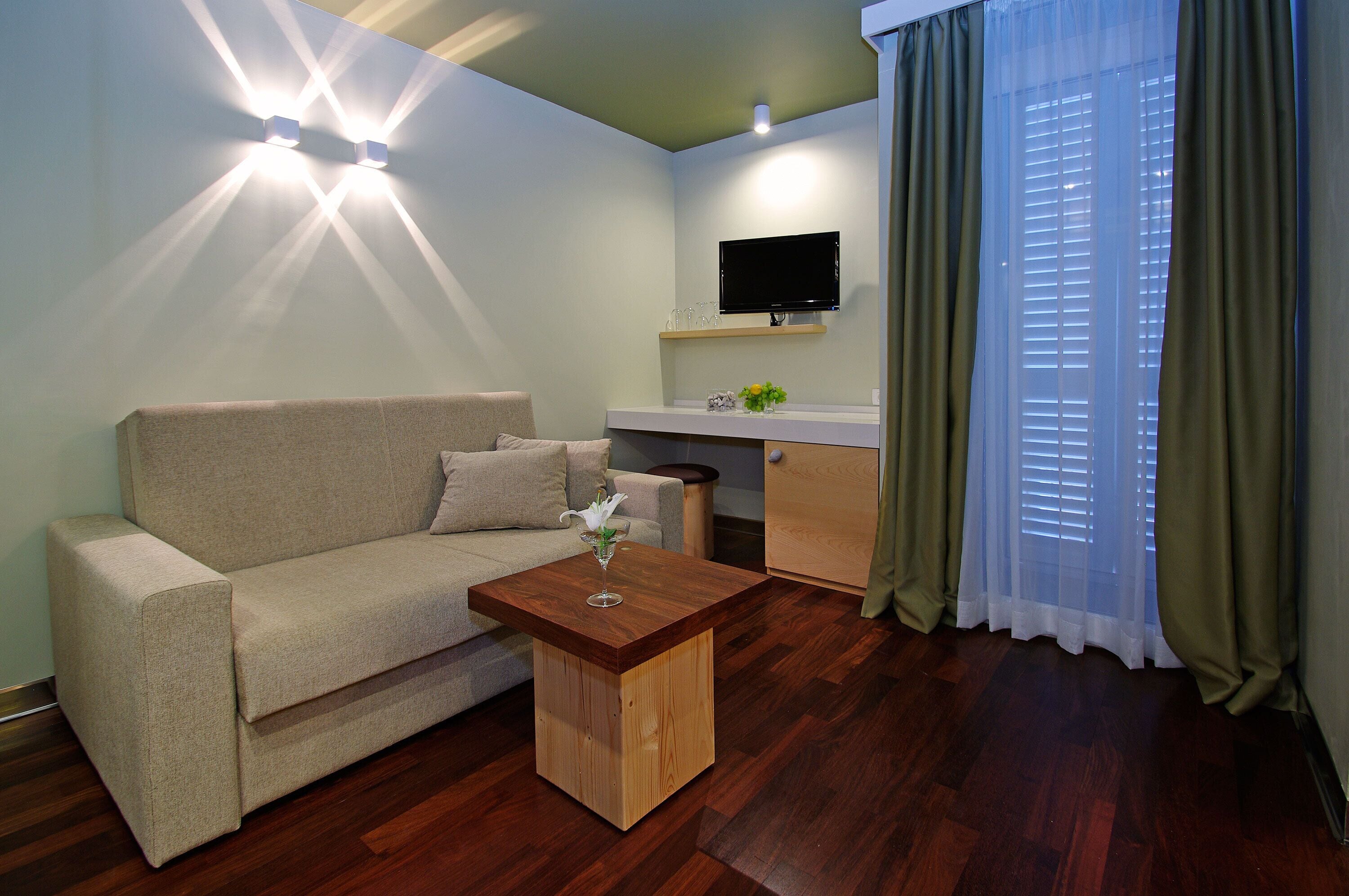 A living room with a sofa, a wooden coffee table, a flat-screen TV mounted on the wall, and a window with curtains.