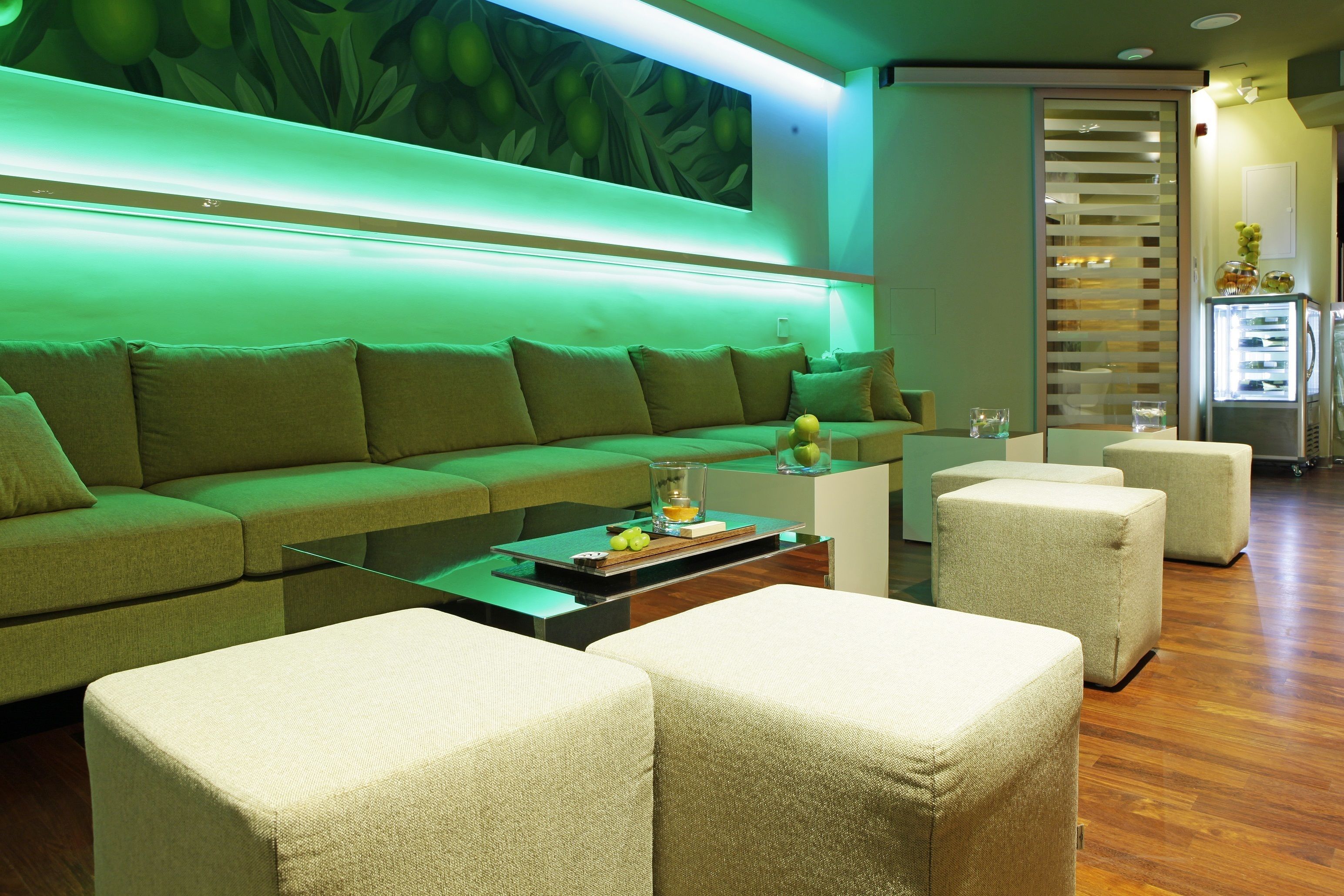 A modern living room with a green-lit wall, a sofa, ottomans, and a coffee table with fruits and a glass.