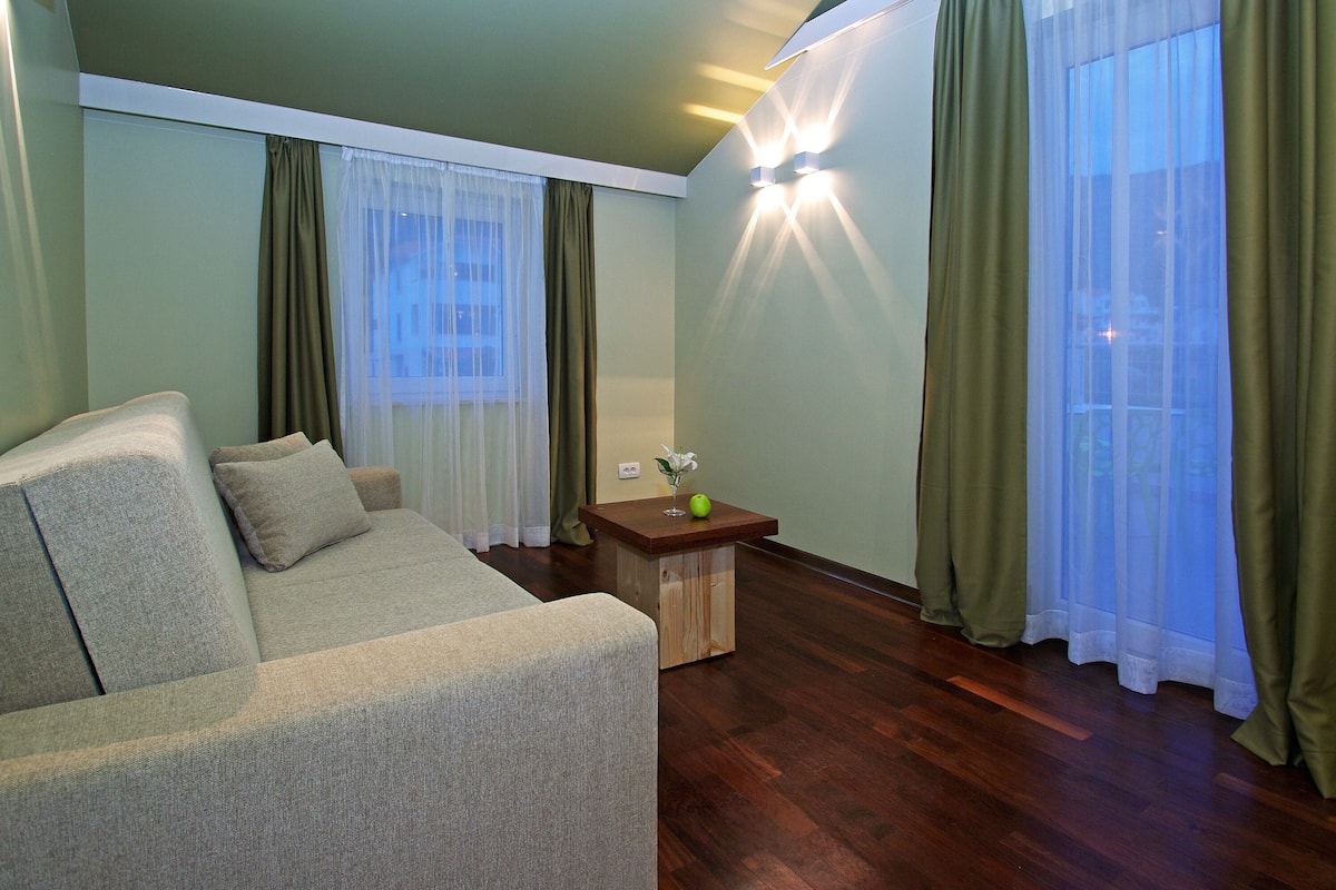 A living room with a sofa, a small wooden table with a vase and an apple, and sheer curtains on the windows.