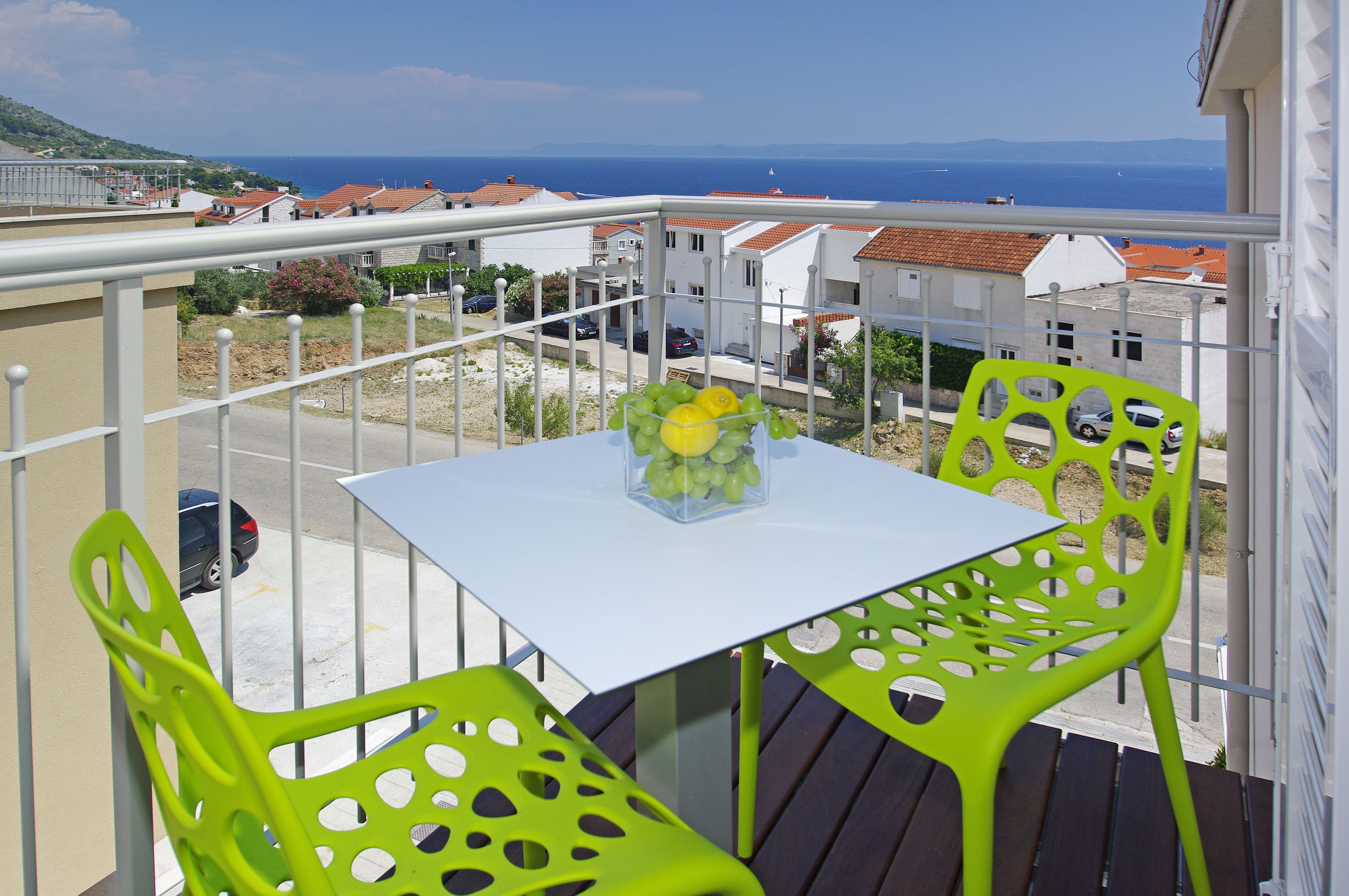 Un balcón con una mesa blanca y dos sillas verdes, con vista a un pueblo costero con casas de tejados rojos y un mar azul cristalino.
