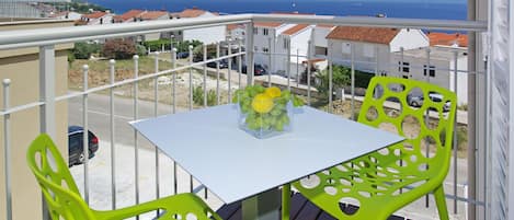 Een balkon met een witte tafel en twee groene stoelen, met uitzicht op een kustplaatsje met rode pannendaken en een heldere blauwe zee.