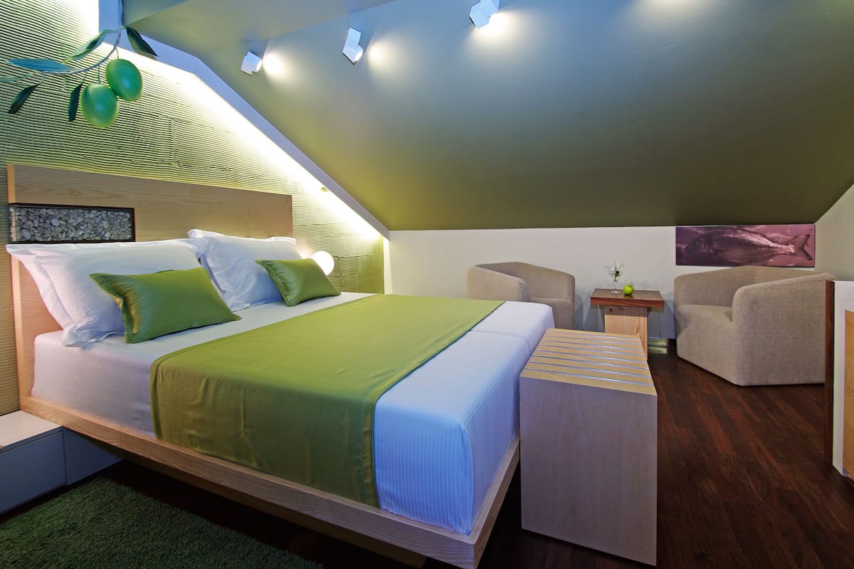 A modern bedroom with a loft-style bed, green bedding, a small seating area, and a painting on the wall.
