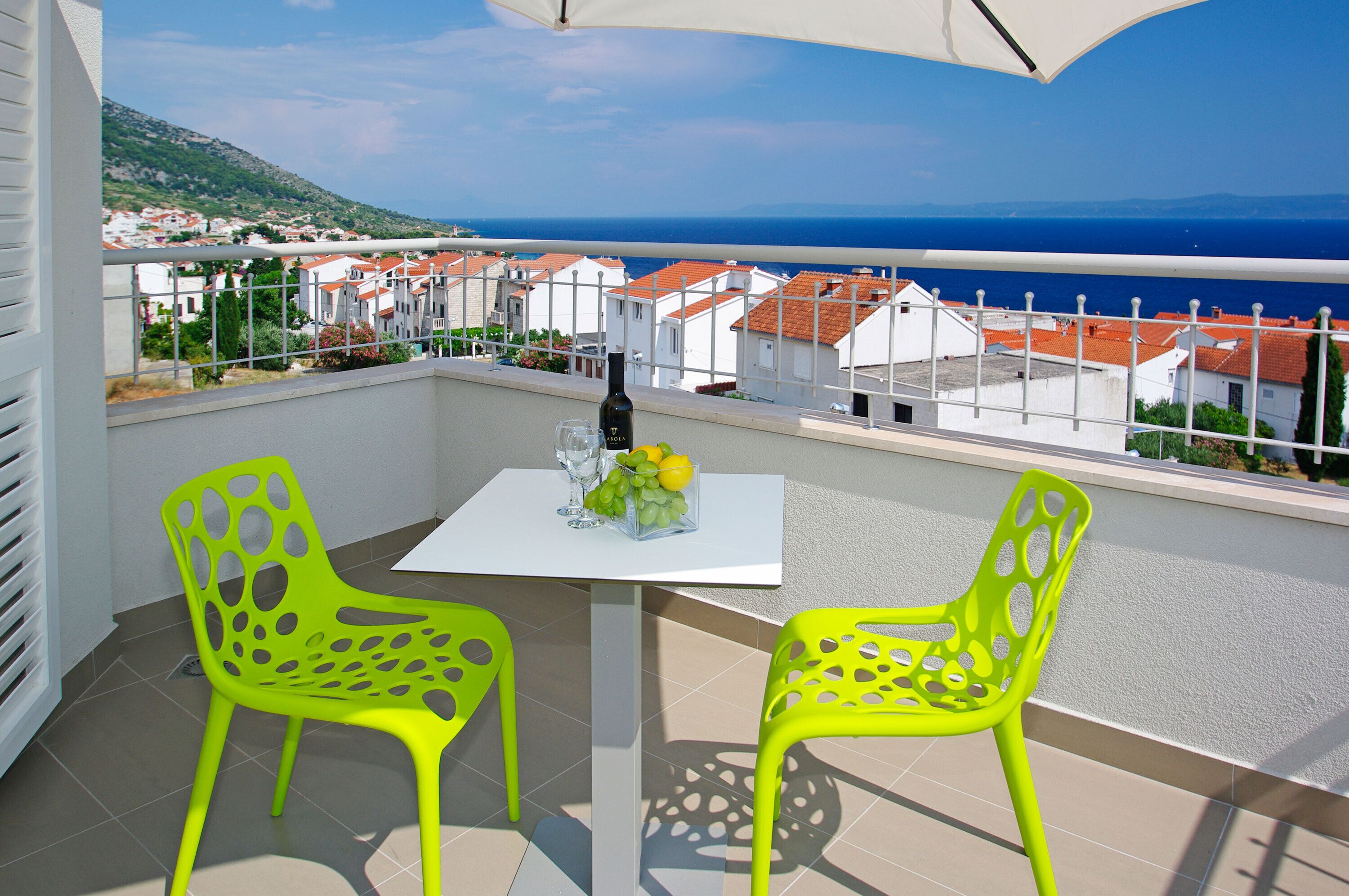A balcony with two bright yellow chairs, a table with a bottle, glasses, and fruit, and a view of a coastal town.