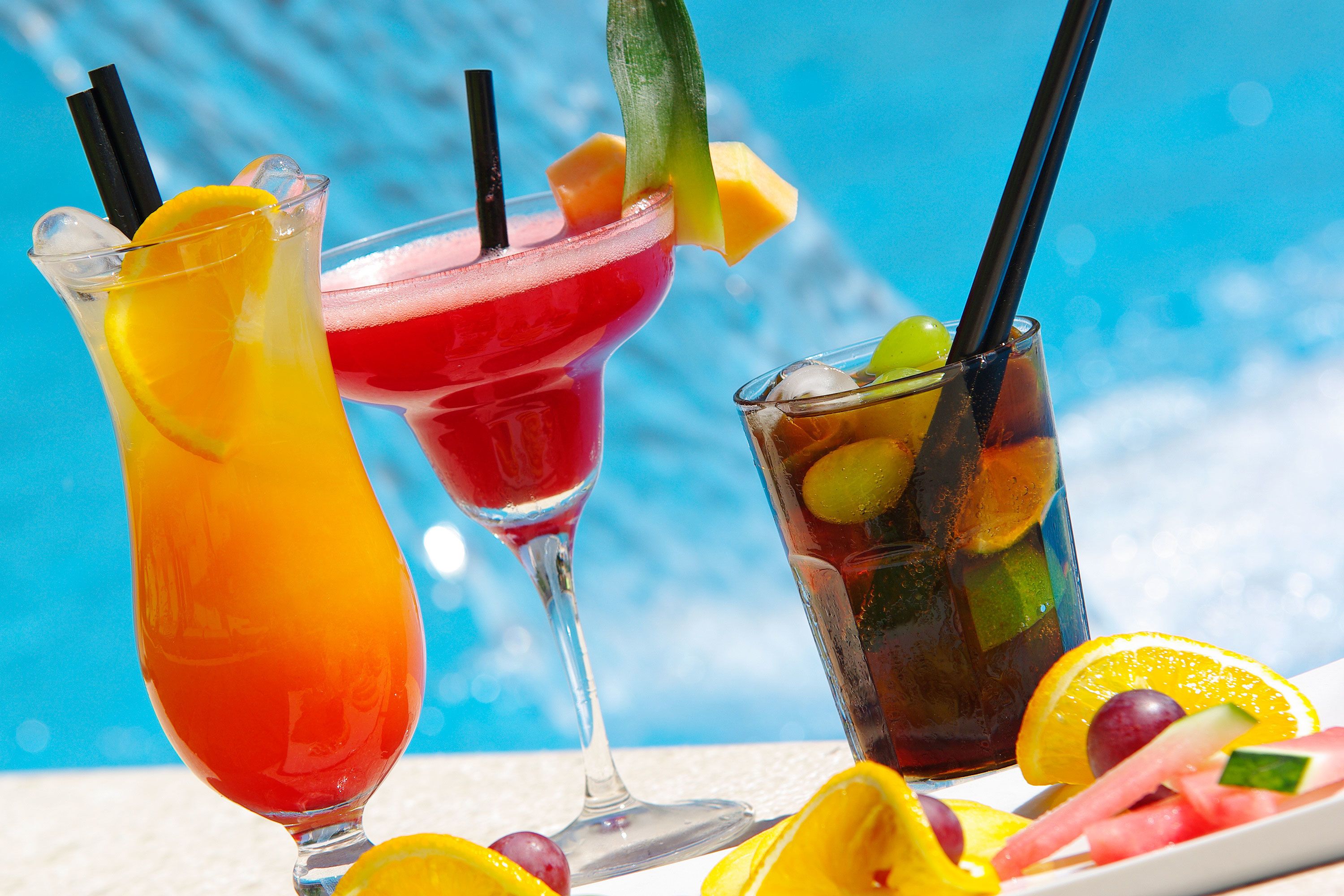 Three different drinks with fruit garnishes and straws, placed on a table with a pool in the background.