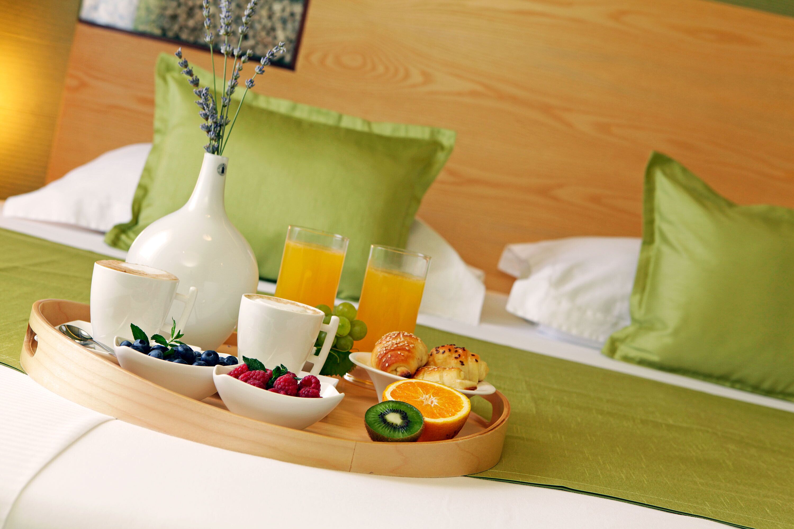 Ein Frühstücksbrett mit Kaffeetassen, Saft, Gebäck und frischem Obst auf einem Bett mit grünen Kissen.