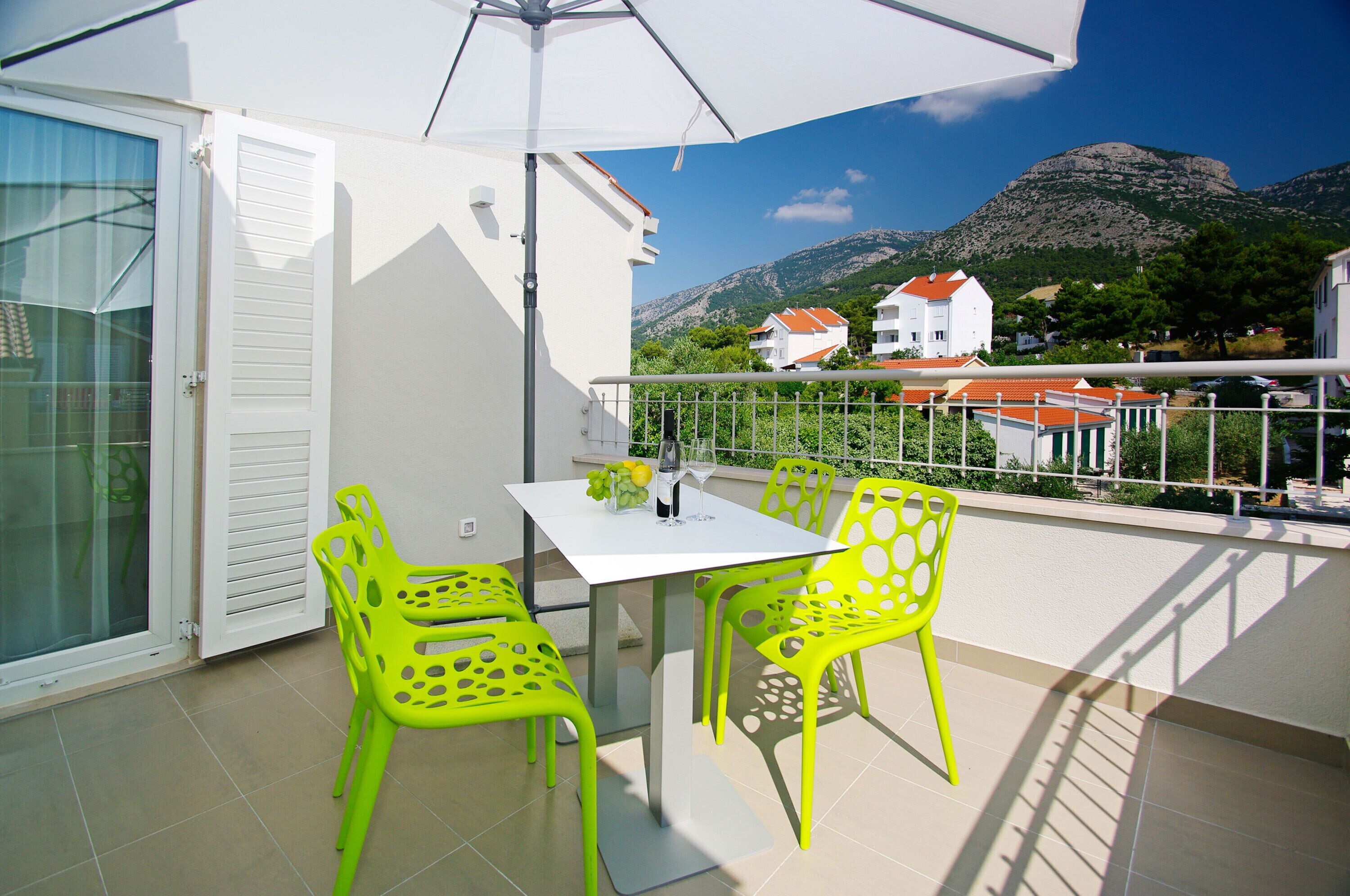 A balcony with a white table and two bright green chairs, a white umbrella, and a view of houses and mountains in the background.