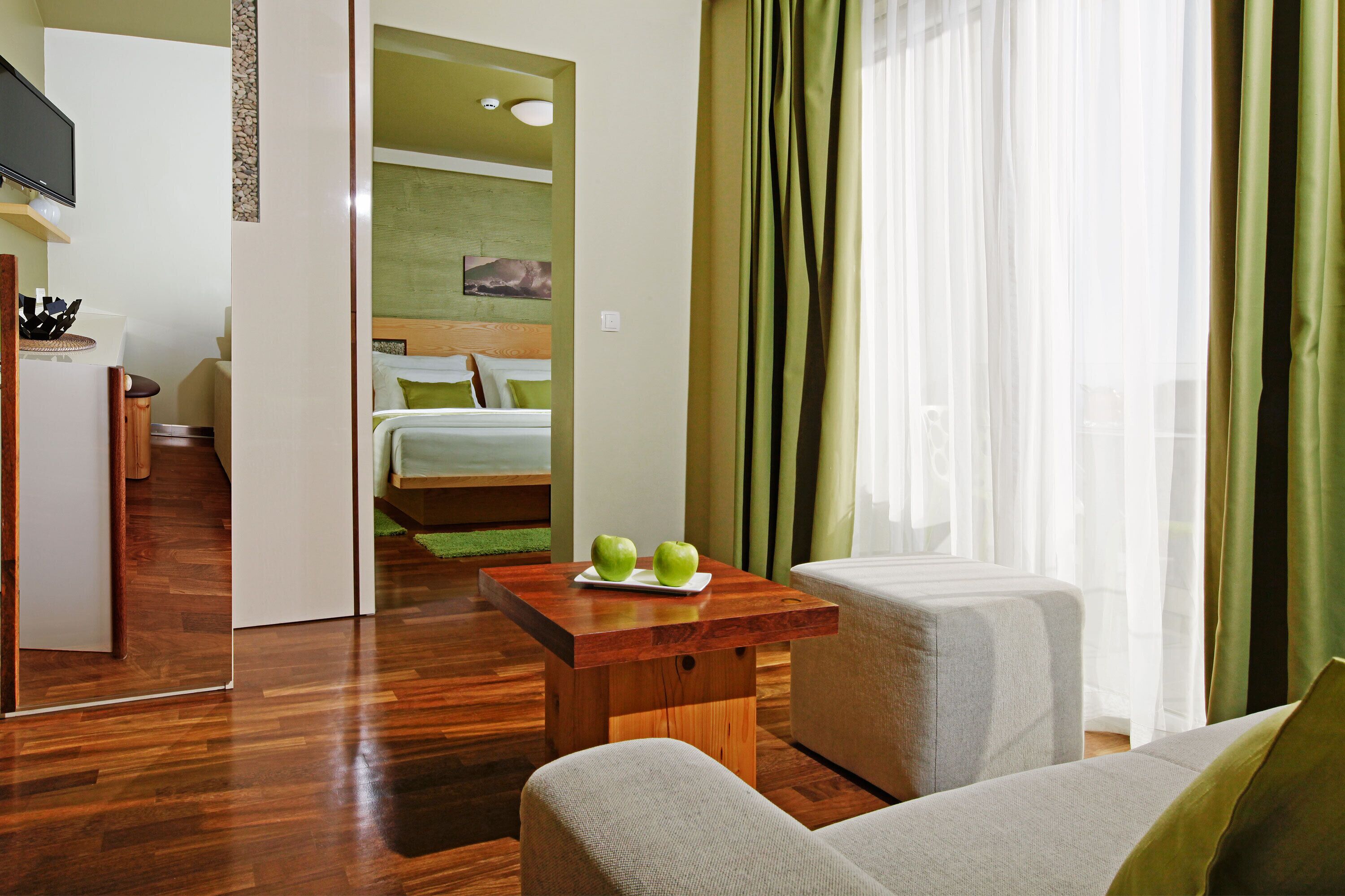 Ein Hotelzimmer mit Holzboden, einer Couch, einem kleinen Tisch mit Äpfeln, einem Bett mit türkisblau-grüner Bettwäsche und Blick auf die Stadt durch transparente Vorhänge.