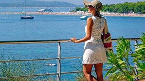 A woman in a white dress and hat standing by a railing overlooking a coastal area with boats and a beach in the background. - Boutique Hotel Bol (Bol)