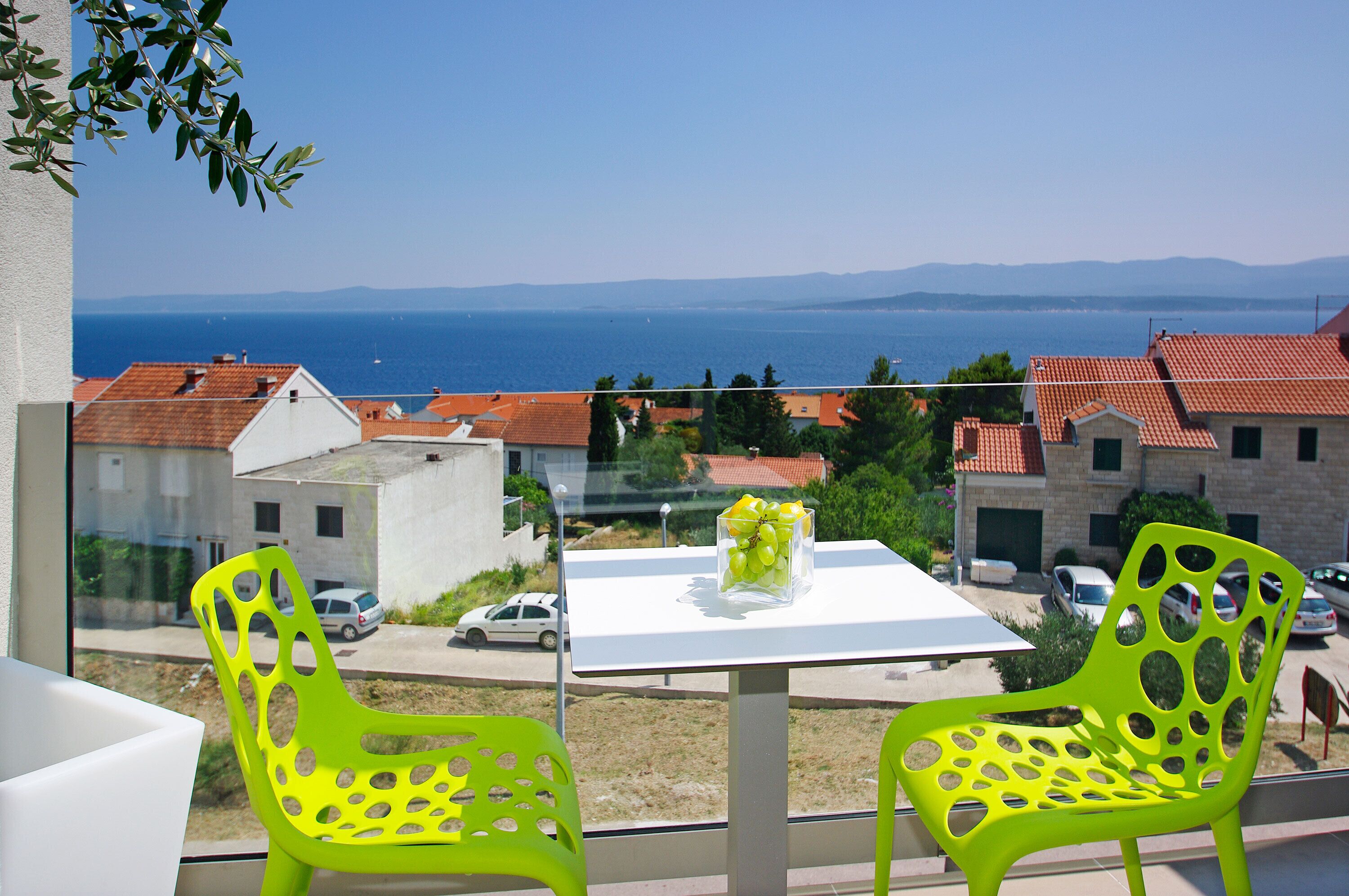 Un balcon avec deux chaises vertes, une table ornée d’un vase de raisins, et une vue sur la mer et les bâtiments.