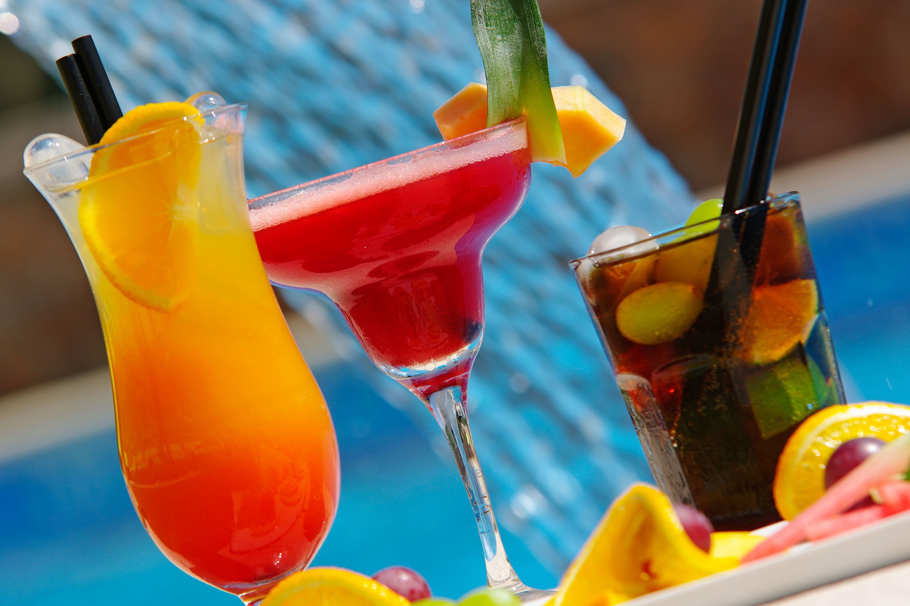 Three cocktails with fruit garnishes and straws, placed on a table by a pool.