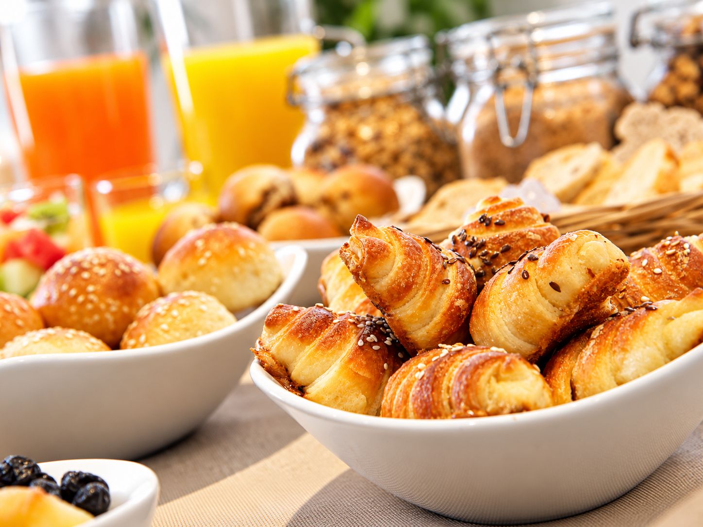 Buffet de café da manhã com diversos pães e doces, incluindo croissants e pães de enrolar, e um copo de suco de laranja.