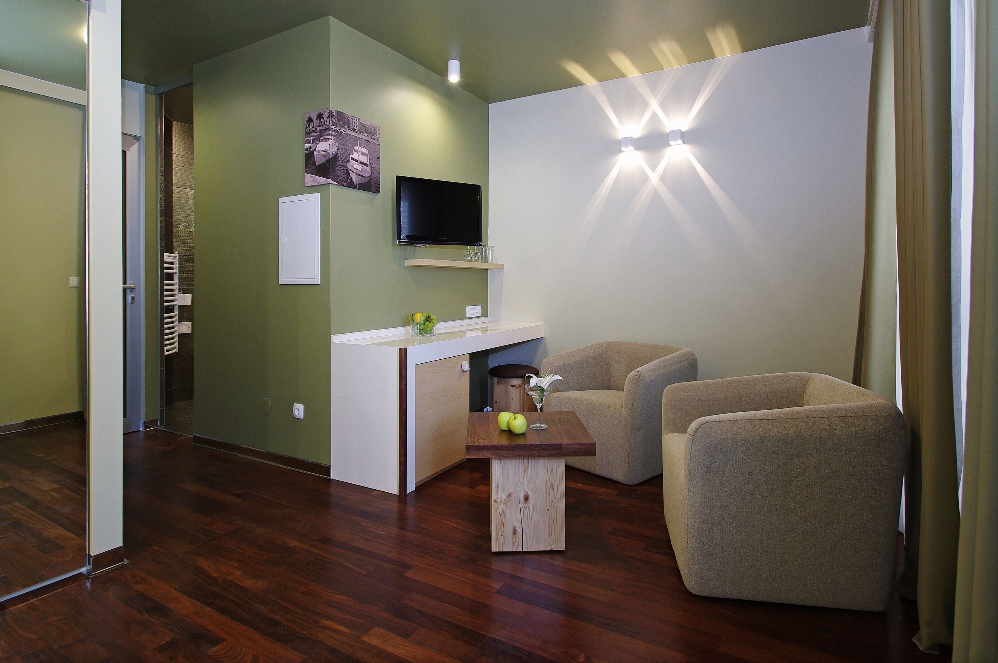 Een woonkamer met een houten salontafel, twee fauteuils, een aan de muur bevestigde televisie en een klein kastje met glazen.