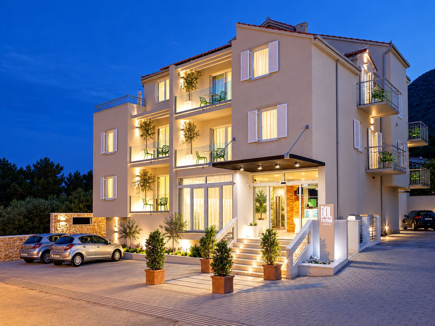 Ein mehrstöckiges Hotel mit einem Balkon im zweiten Stock, einer Treppe, die zum Eingang führt, und einem Parkplatz mit Autos.