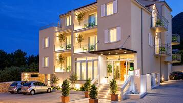 Ein mehrstöckiges Hotel mit einem Balkon im zweiten Stock, einer Treppe, die zum Eingang führt, und einem Parkplatz mit Autos.