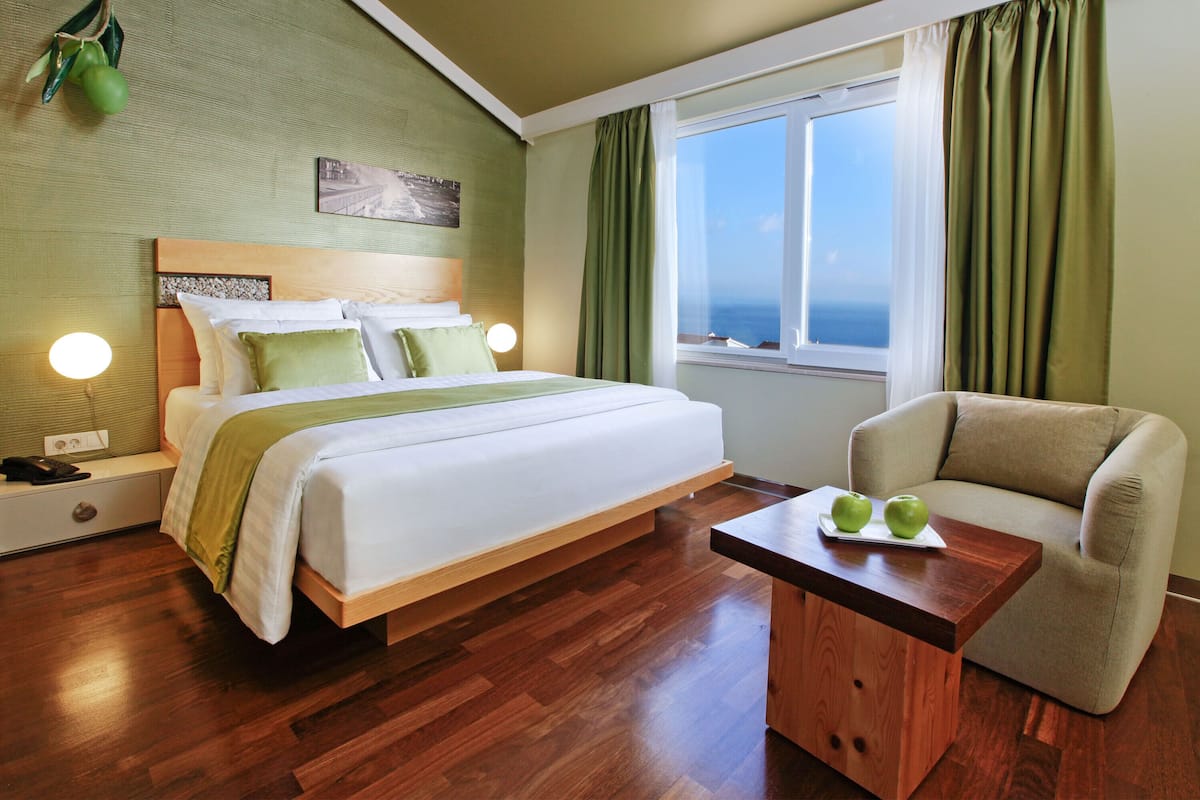 A hotel room with a bed, a wooden nightstand, a table with apples, a chair, and a window with curtains.