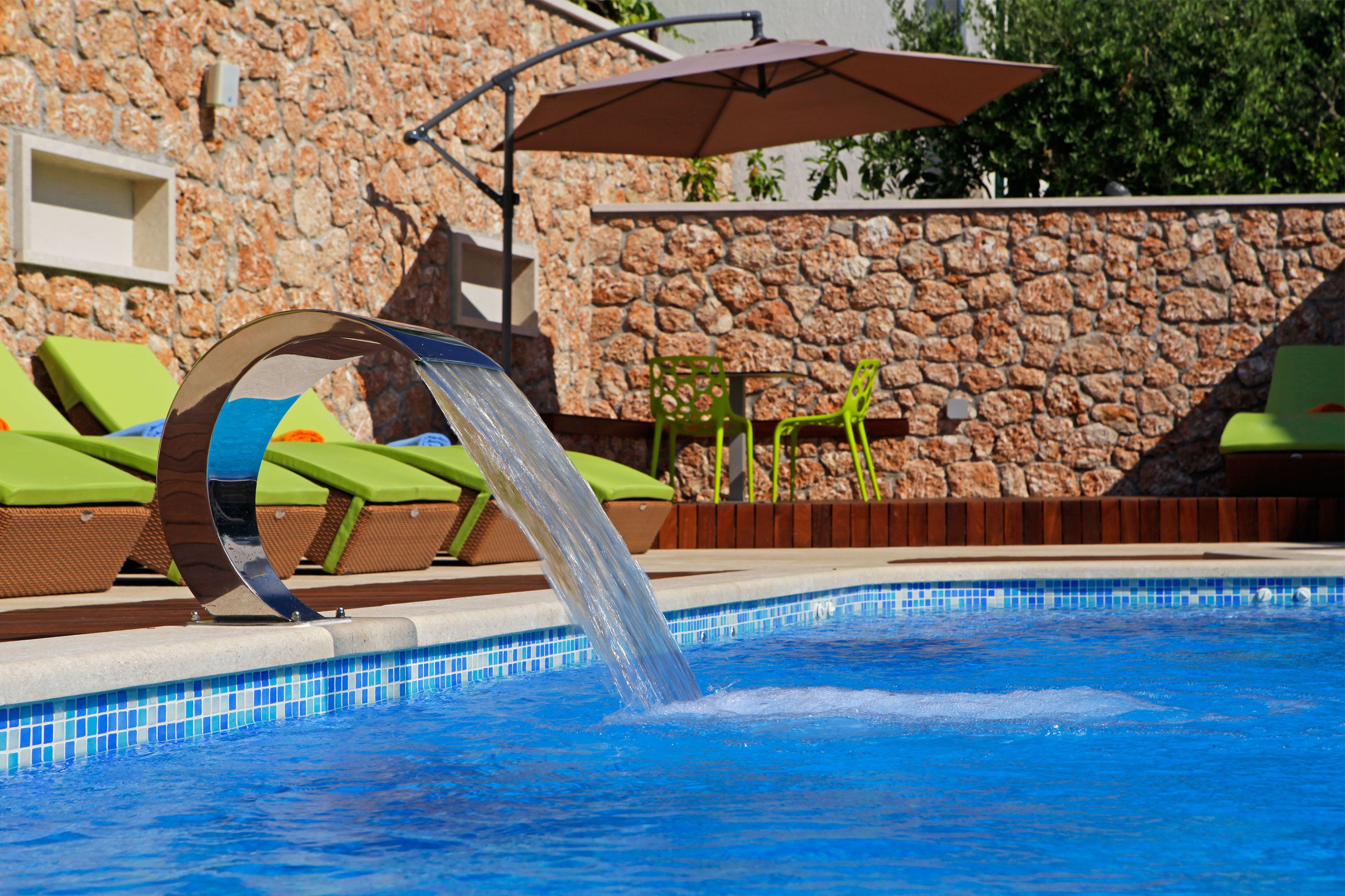 Une piscine agrémentée d’un élément décoratif aquatique, des chaises longues vertes et un mur de pierre en arrière-plan.