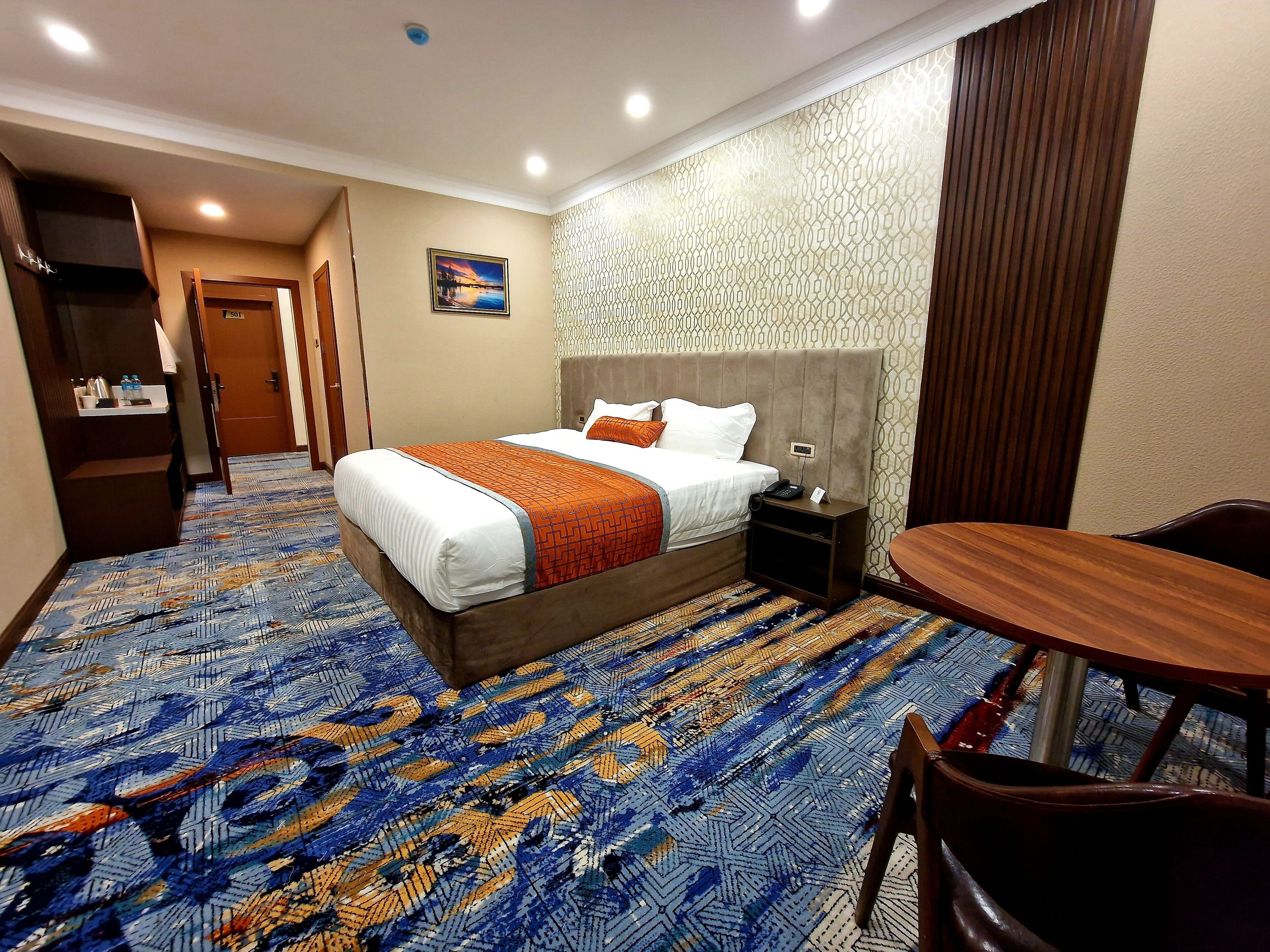 A hotel room with a large bed, a bedside table, a chair, a small table, and a patterned carpet.