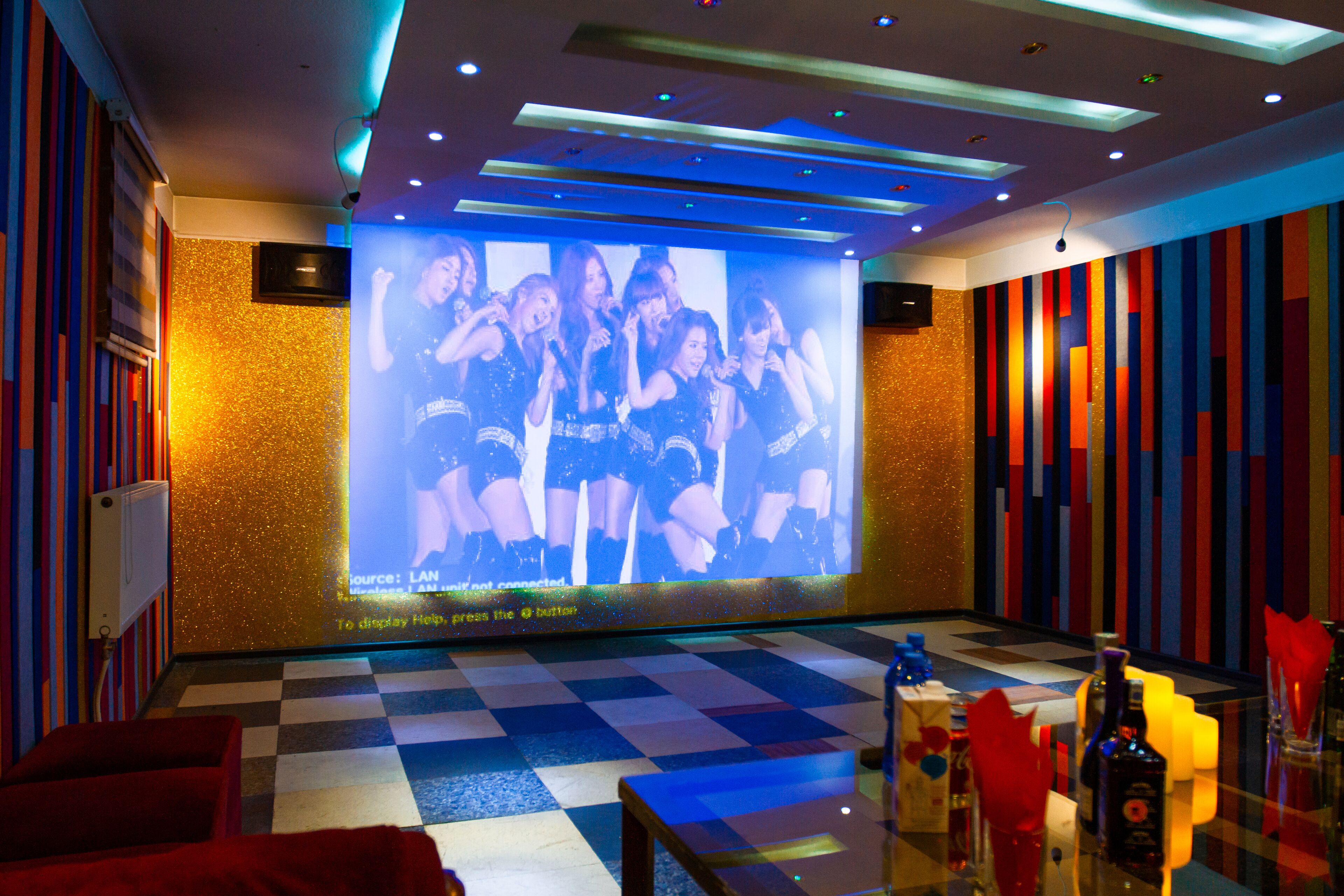 Habitación de hotel con una pantalla grande mostrando un grupo de personas, suelo a cuadros y paredes con rayas de colores.