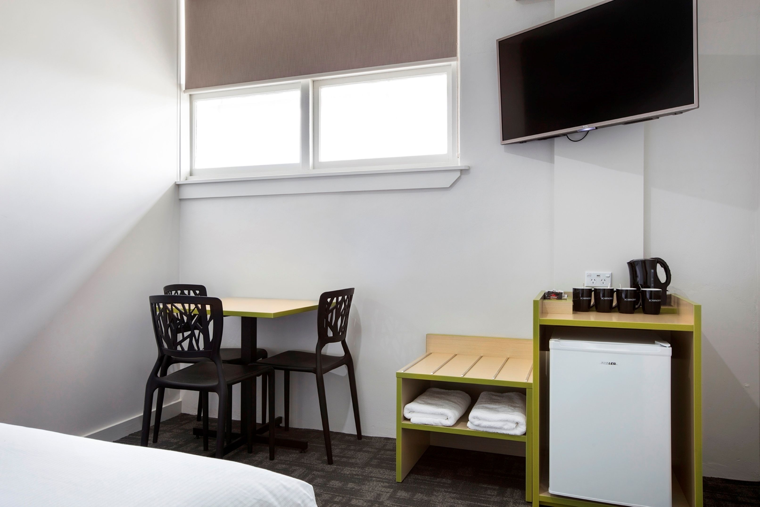 A hotel room with a bed, a desk with two chairs, a TV mounted on the wall, a small refrigerator, and a shelf with towels and cups.