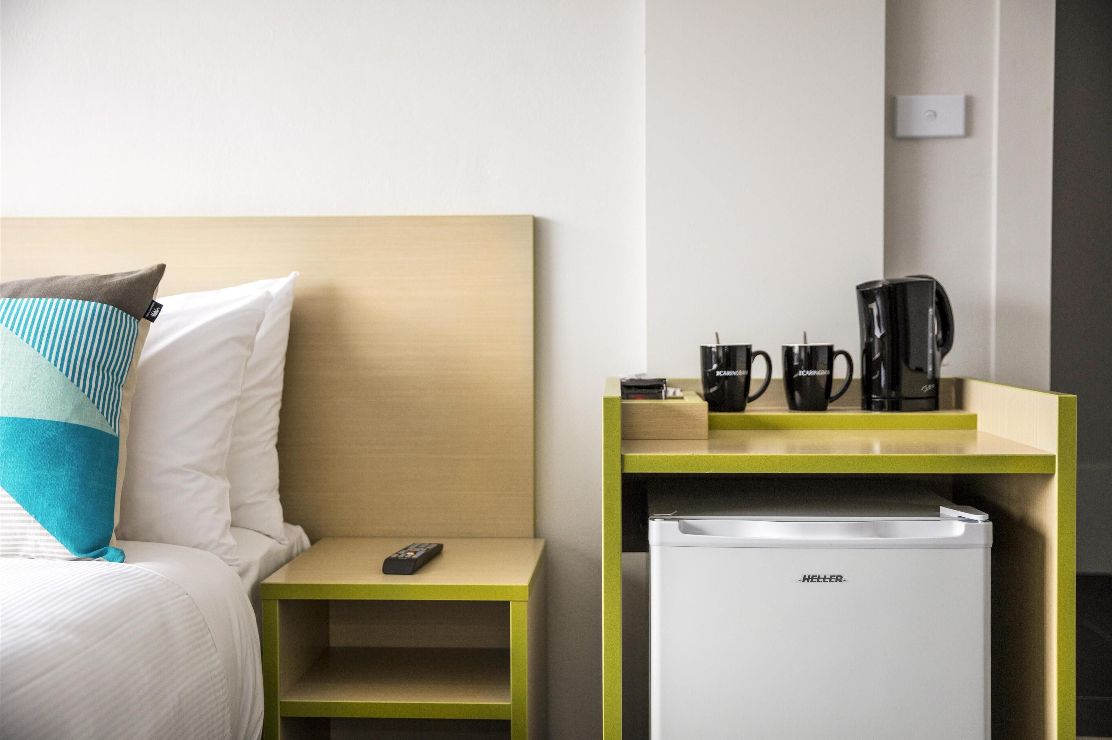 Una habitación de hotel con una cama, una mesita de noche con control remoto, una cafetera y un refrigerador.