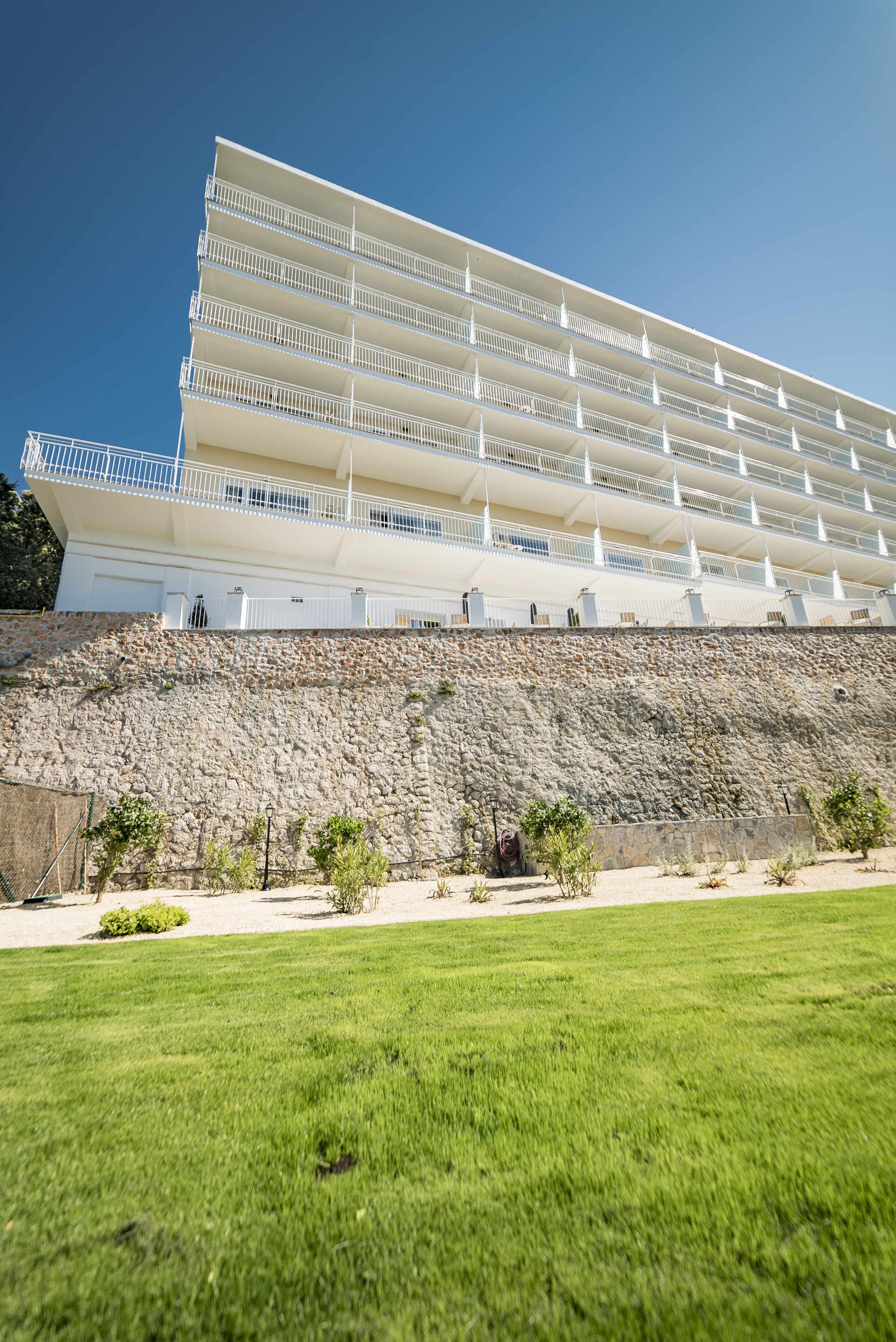 A multi-story hotel with balconies, a stone retaining wall, and a grassy lawn.