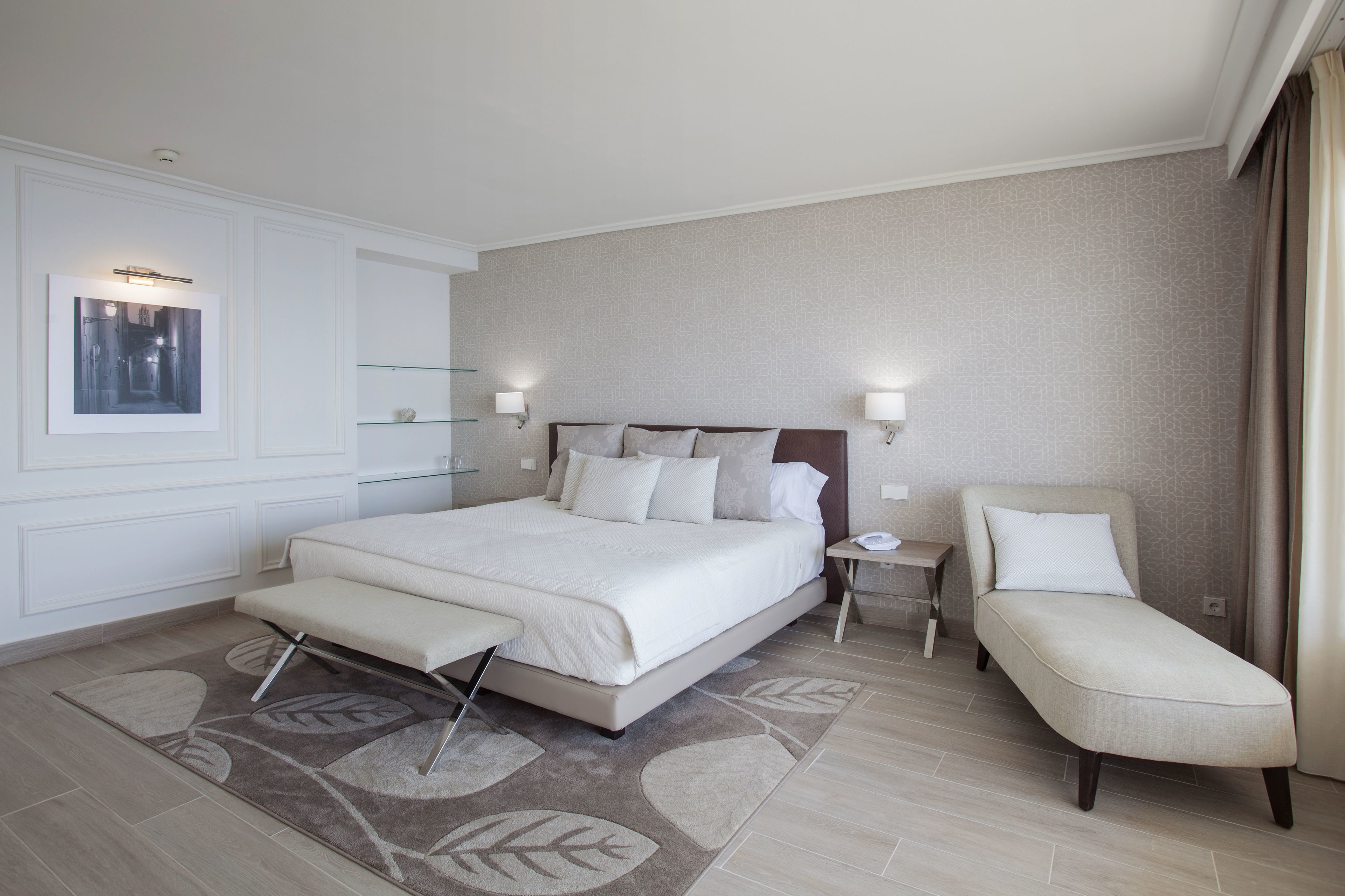 A modern hotel room with a large bed, a bedside table, a chair, and a small table with a lamp.
