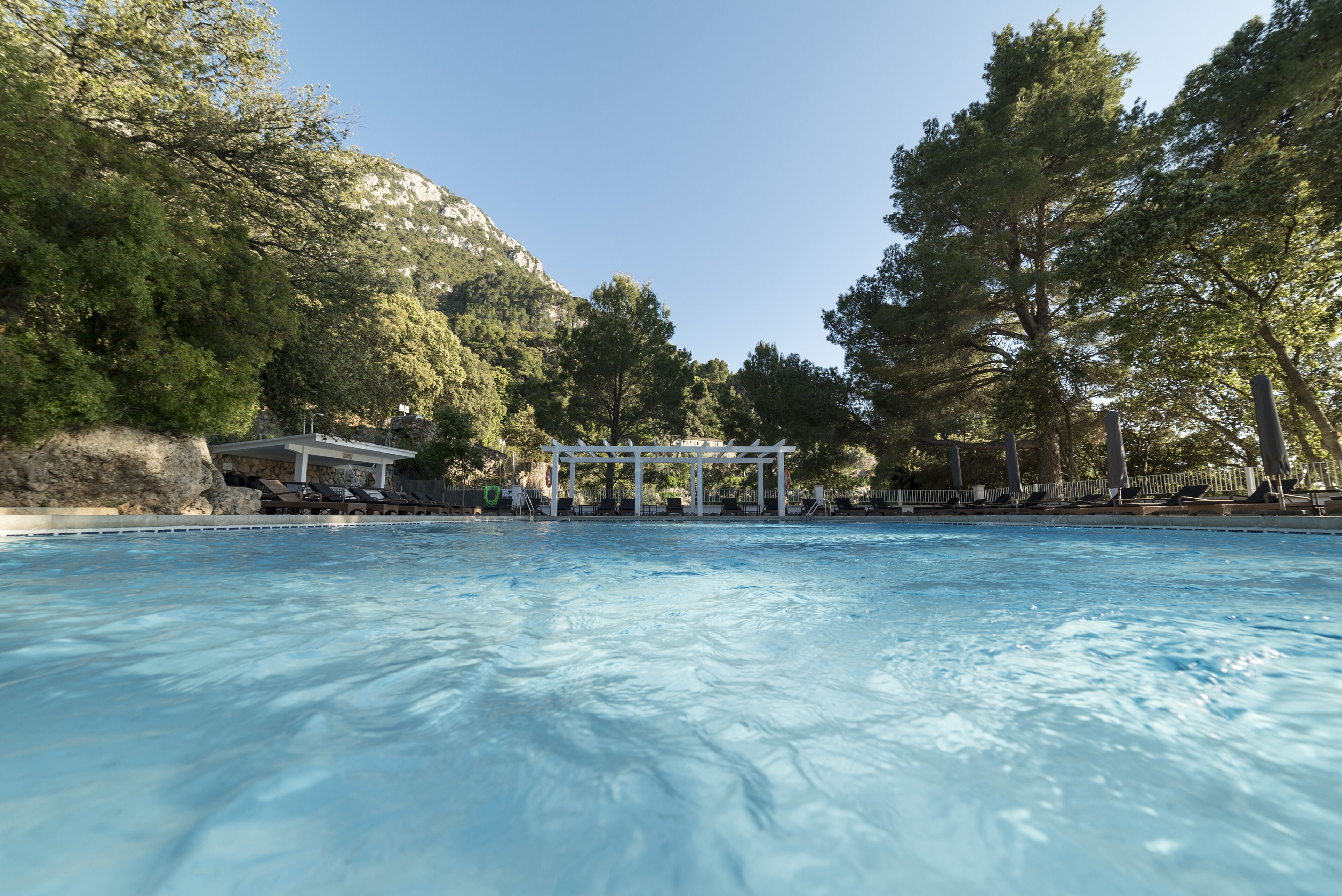 A swimming pool with a gazebo and lounge chairs, surrounded by trees and a mountain backdrop.