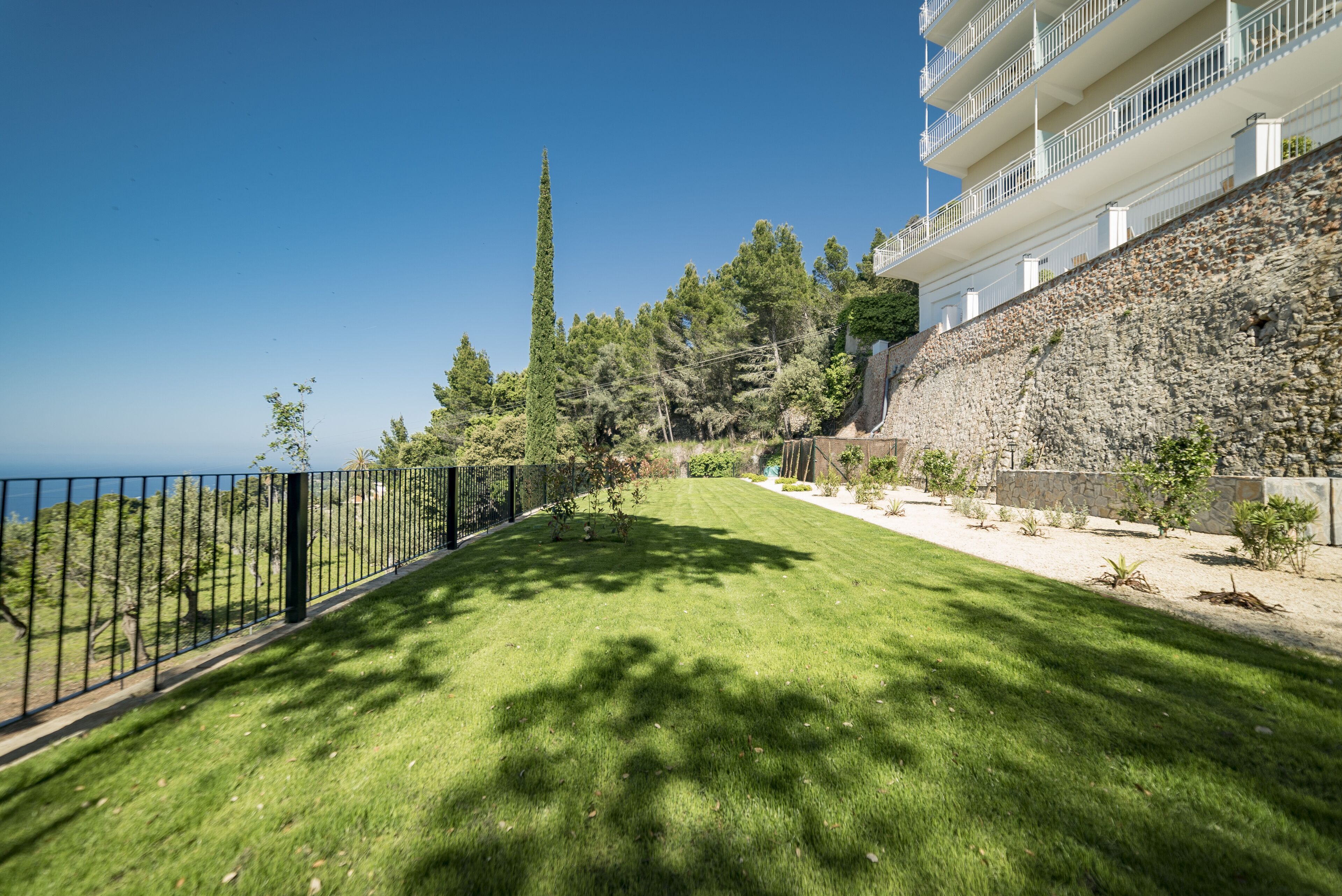 A well-maintained lawn with a stone retaining wall, a black metal fence, and a building with balconies overlooking a body of water.