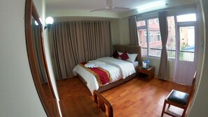 A hotel room with a bed, a wooden chair, a nightstand, a ceiling fan, and a view of buildings through the window. - Om Tara Guest House (Kathmandu)