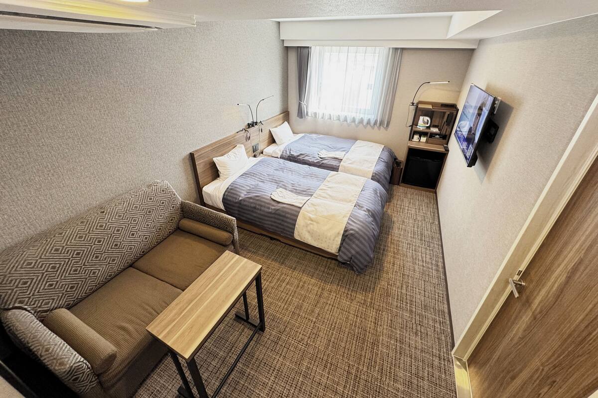 A hotel room with two beds, a sofa, a small table, a TV, and a window with curtains.
