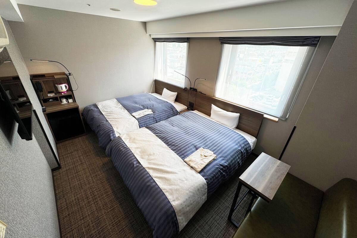 A hotel room with two beds, a desk, a chair, a coffee maker, and a window with blinds.