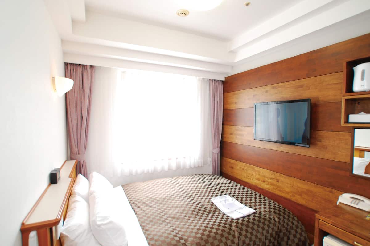 A hotel room with a large bed, a television mounted on the wooden wall, a bedside table with a lamp, and a window with curtains.