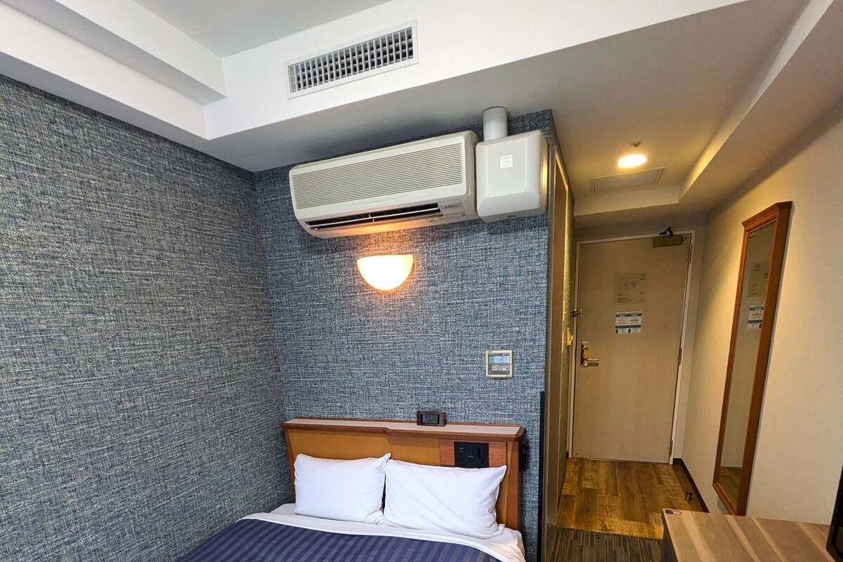 A hotel room with a bed, a desk, an air conditioner, a wall-mounted light, and a door leading to another room.