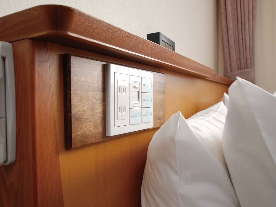 Ein Hotelzimmer mit einem Holzkopfstück, einer Steuerung mit mehreren Tasten und einem weißen Kissen.