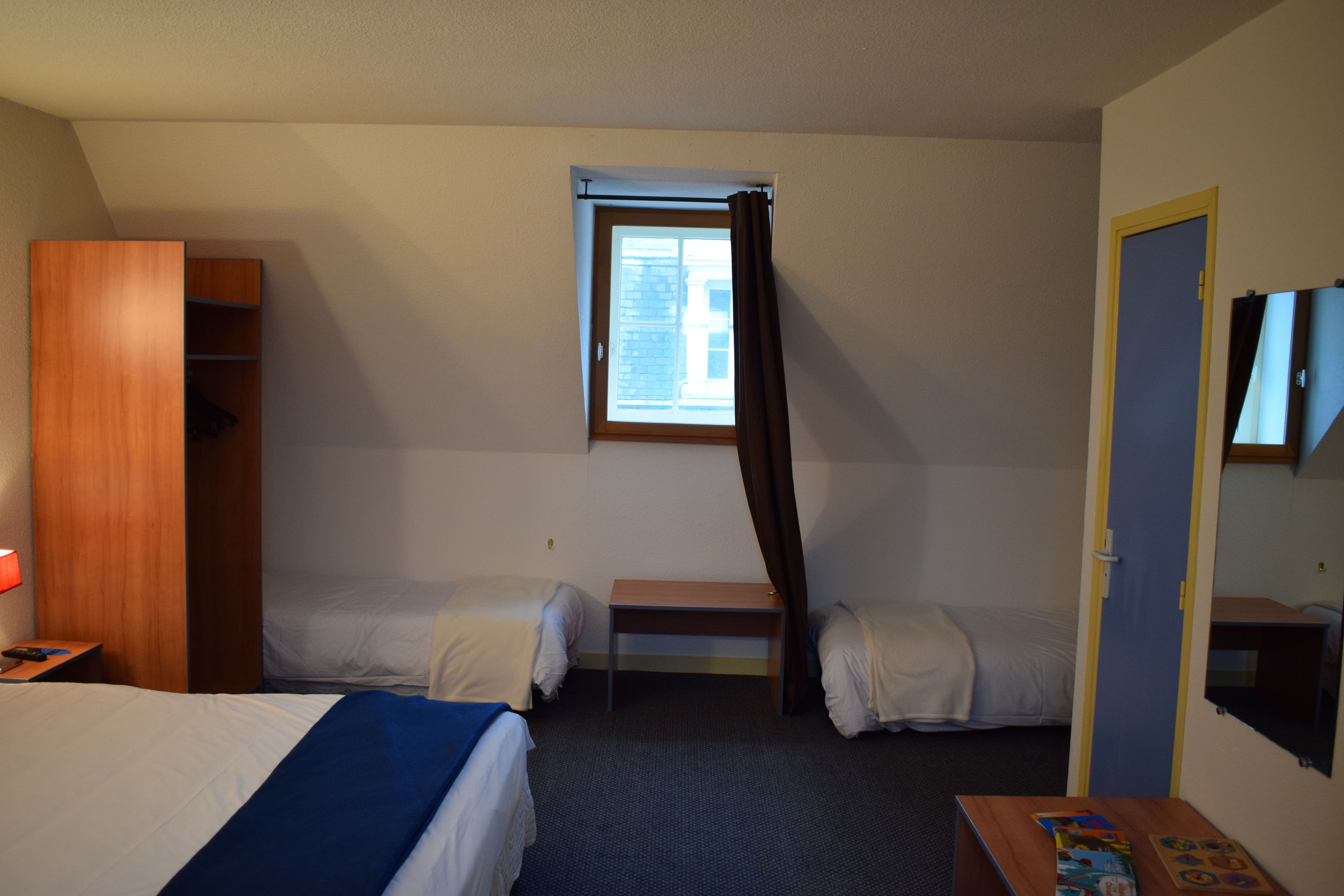 Habitación de hotel con dos camas individuales, un armario de madera, una mesita pequeña, una ventana con cortinas y un televisor en la pared.