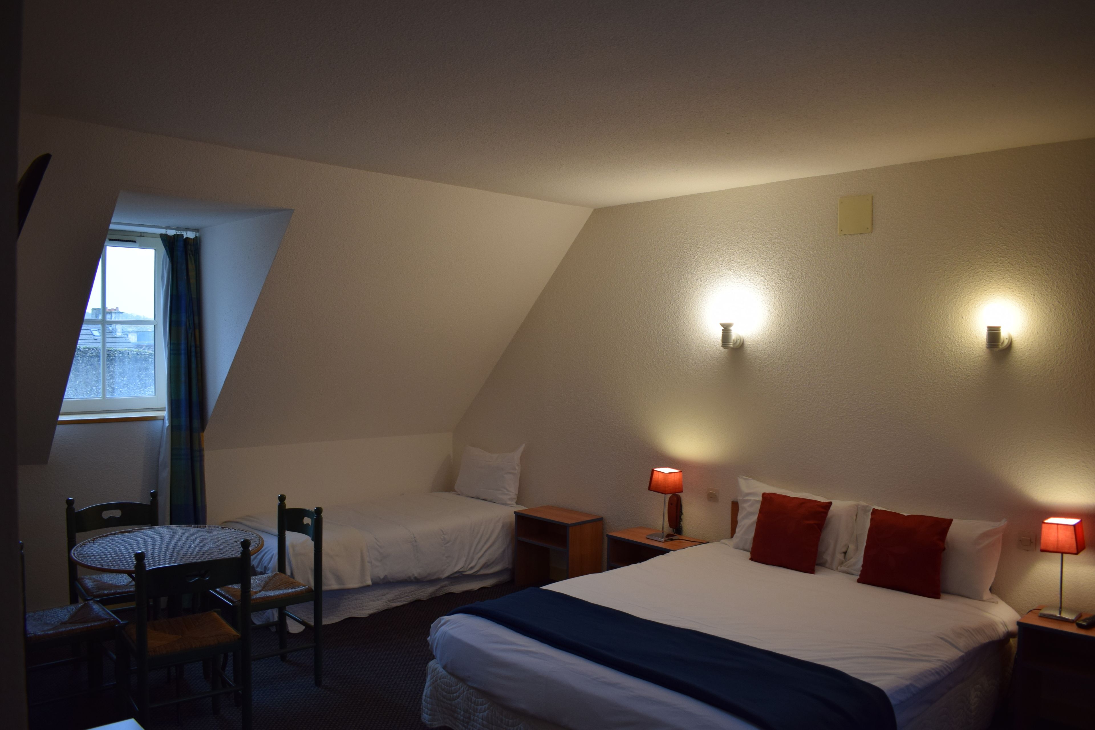 Habitación con dos camas individuales, una mesita pequeña y sillas. Hay dos lámparas de noche y una ventana con cortinas.