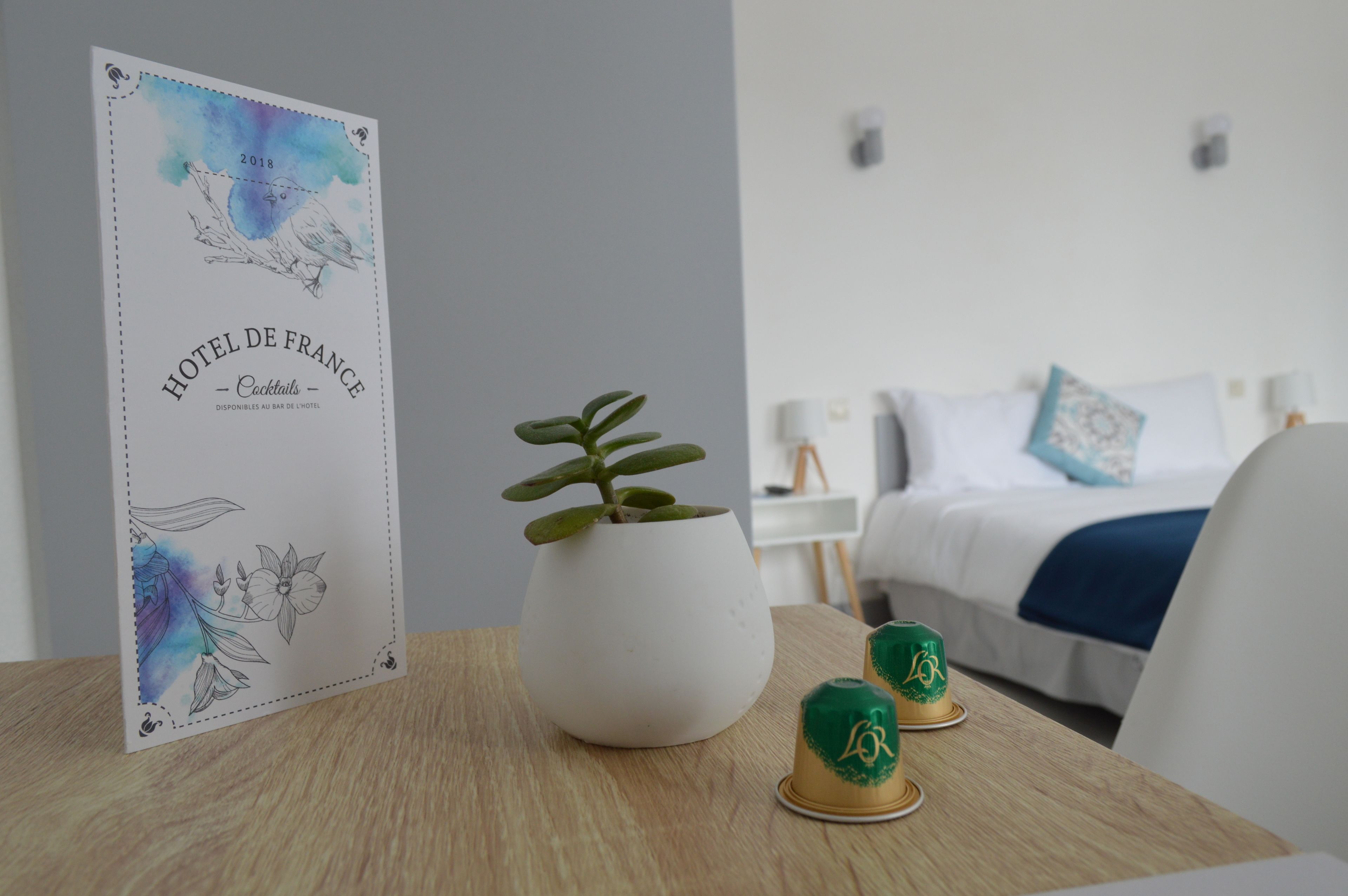 A hotel room with a bed, a bedside table with a plant and a framed menu, and a nightstand with a lamp.