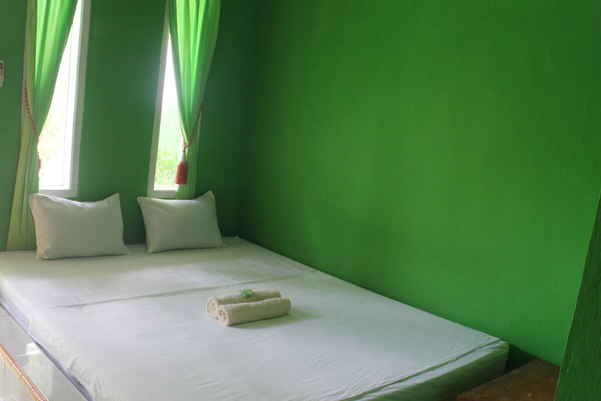 A single bed with white bedding and pillows, a small bedside table, and a window with green curtains.