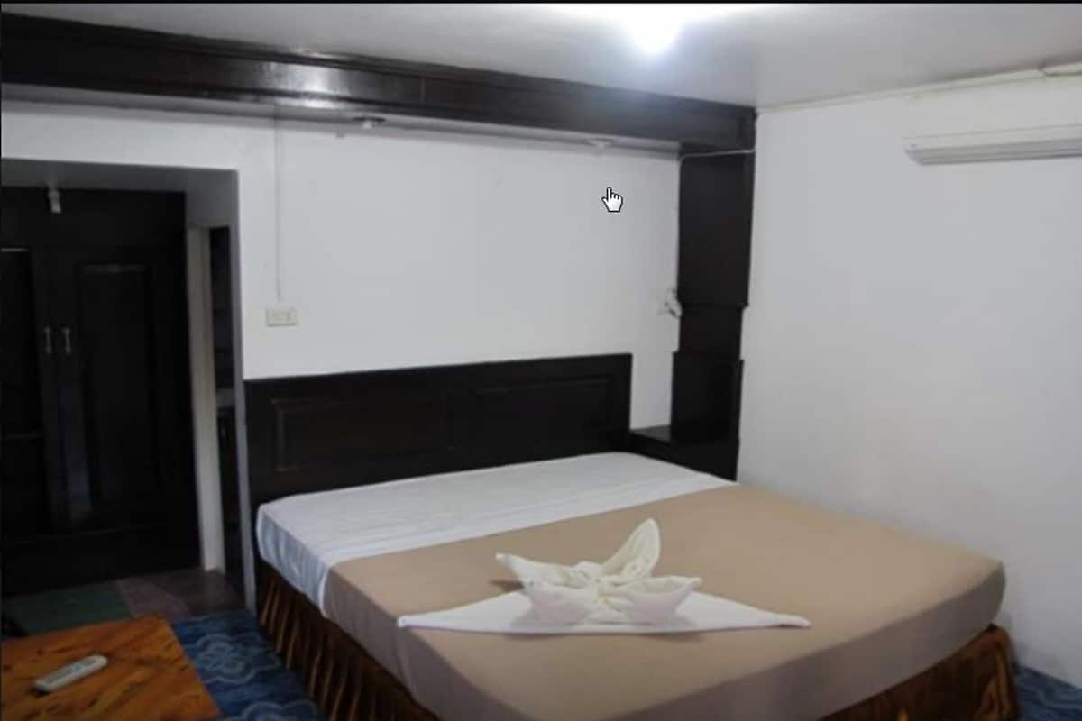 A single bed with a white bedspread and a brown skirt, a wooden nightstand with a remote control, and a door to the left.