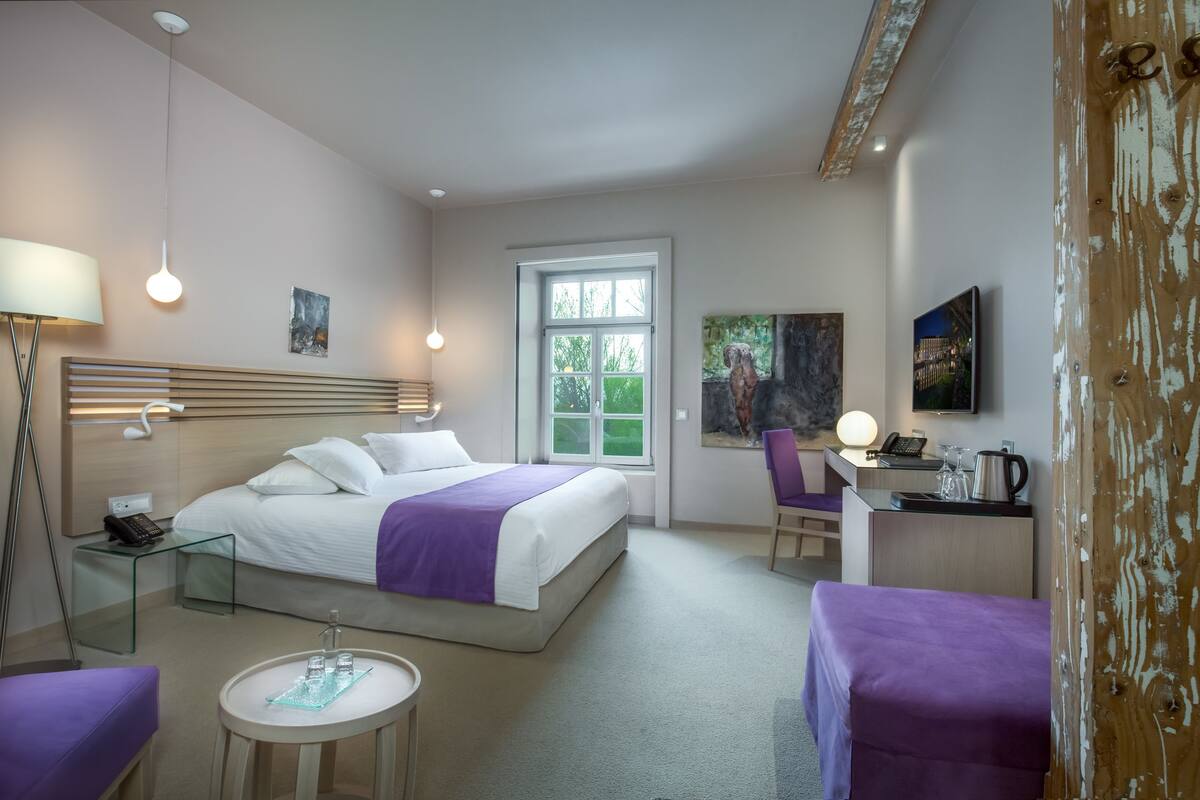 Ein Hotelzimmer mit Bett, Schreibtisch mit Stuhl, einem kleinen Tisch und einem Fenster mit Blick ins Grüne.
