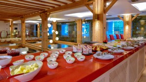 A buffet setup with various dishes and bowls on a red counter, wooden beams on the ceiling, and a view of the room in the background. - Hydrama Grand Hotel (Drama)