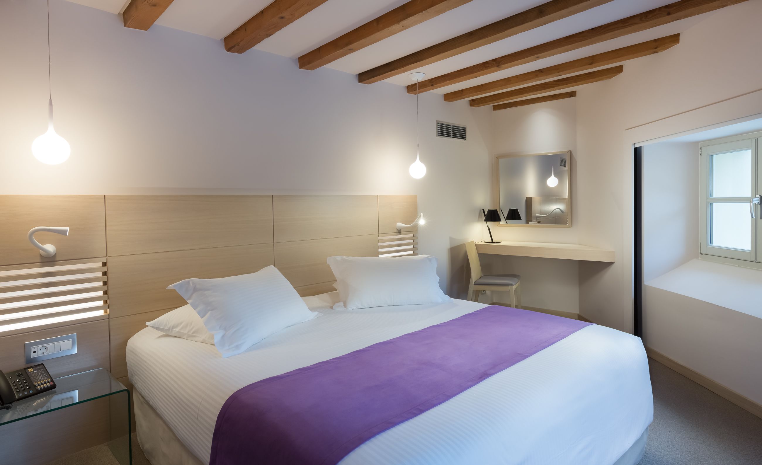 Una habitación de hotel moderna con una cama grande, una mesita de noche con teléfono, un escritorio con silla y una ventana.