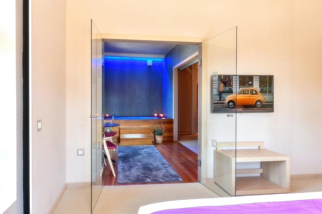 A modern hotel room with a glass door leading to a bathroom, a small seating area with a pool table, and a framed picture on the wall.