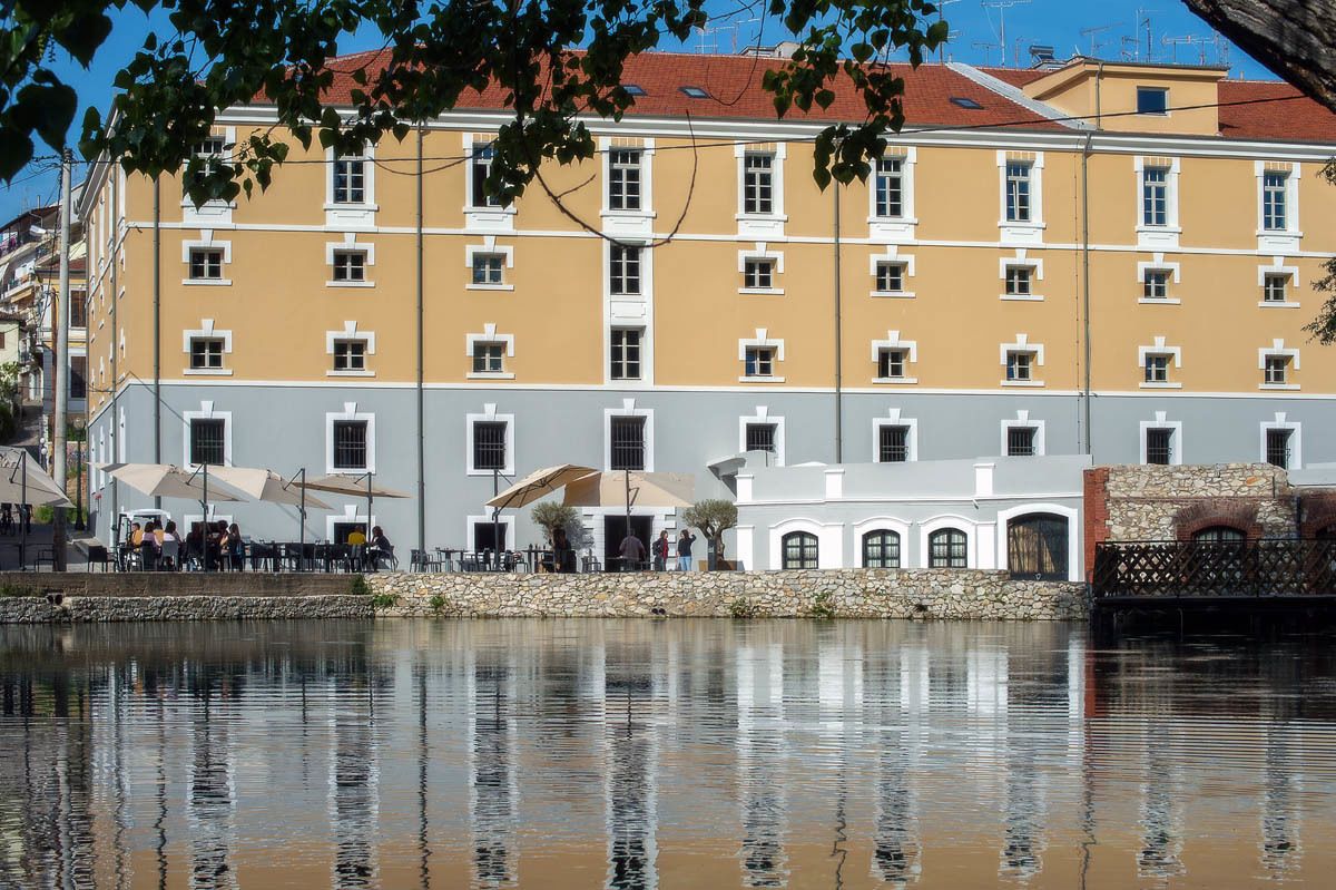 A multi-story building with a yellow facade, white trim, and a stone base. There are outdoor tables and umbrellas, and a body of water in front.