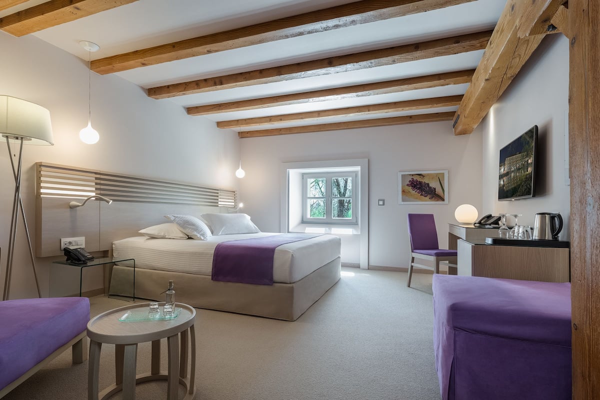 Ein modernes Schlafzimmer mit Bett, Nachttisch, Lampe und einem Fenster mit Blick auf Bäume.