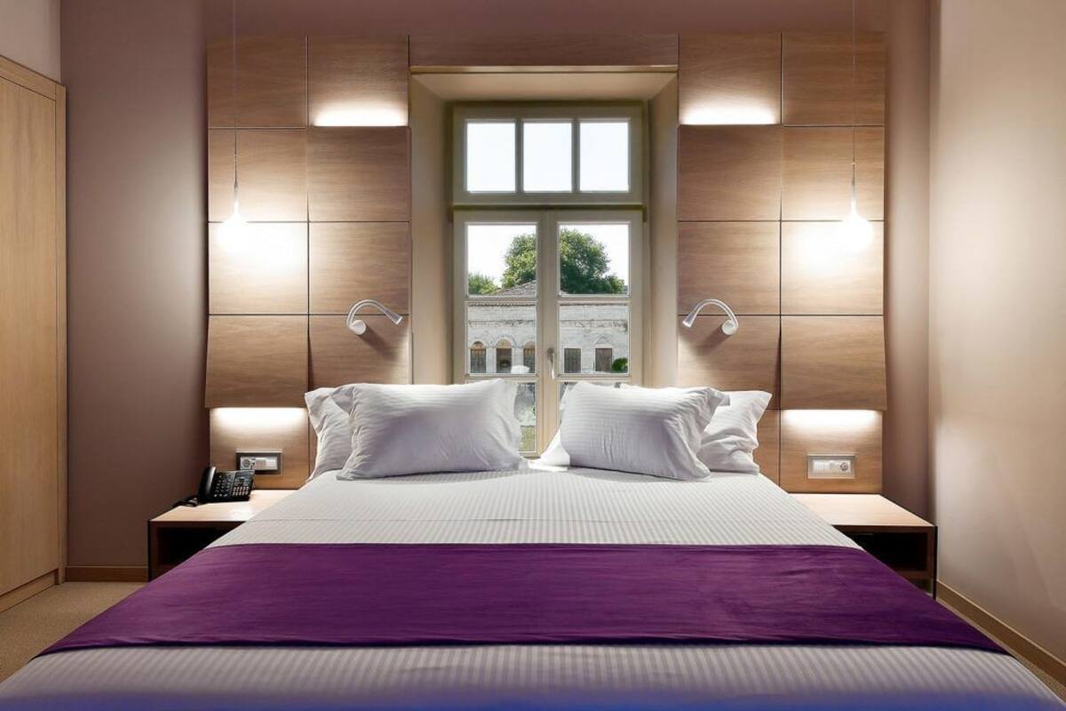 Ein modernes Hotelzimmer mit einem großen Bett, zwei Nachttischen, einem Fenster mit Blick auf ein Gebäude und Wandleuchten.