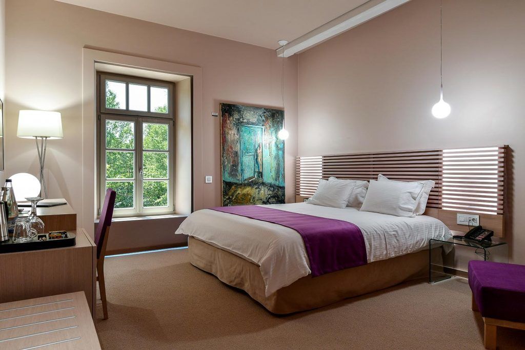 A bedroom with a bed, bedside table, lamp, window, and artwork.