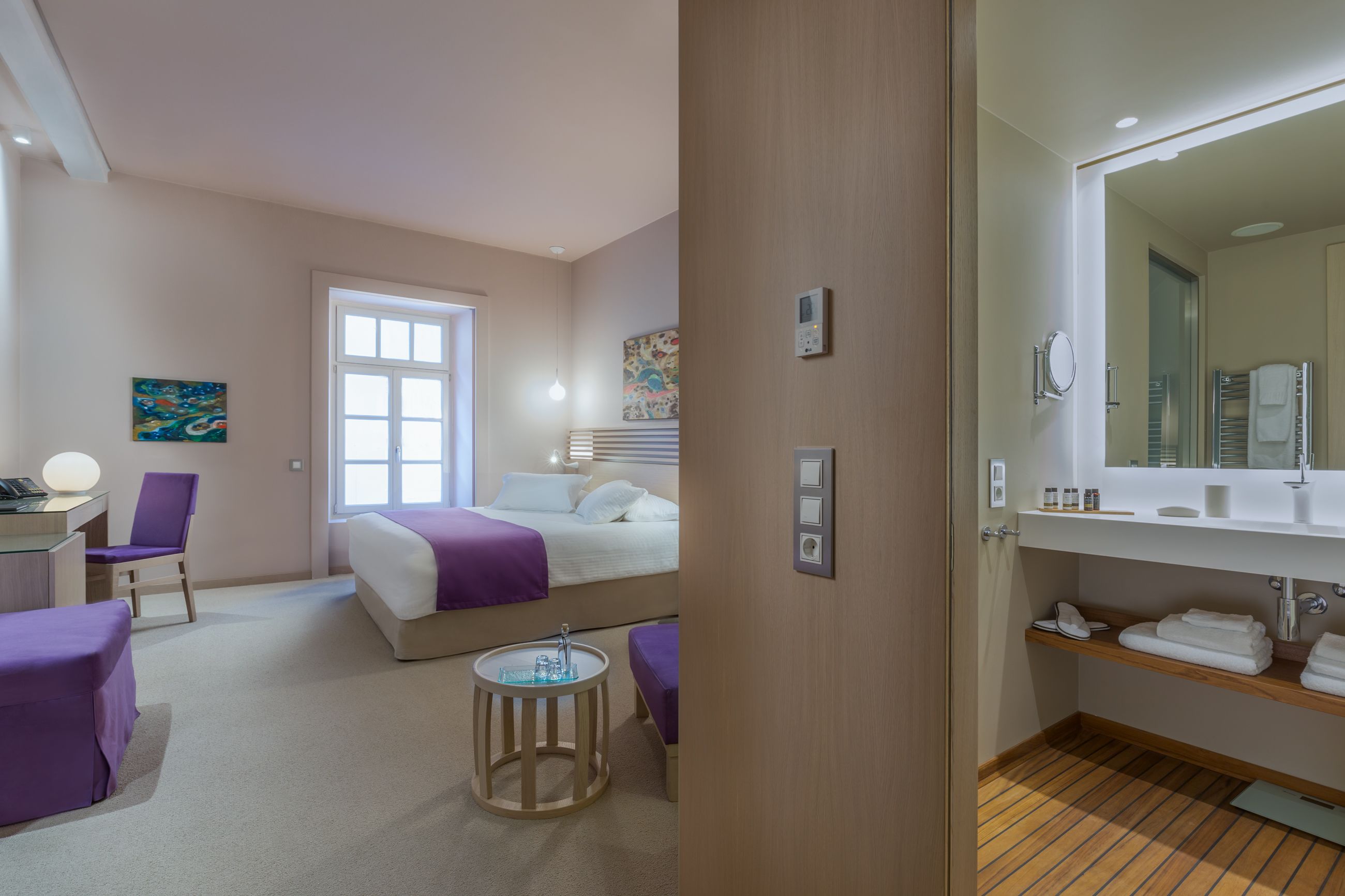Habitación de hotel con cama, escritorio, silla, una mesita redonda y baño con lavabo y espejo.
