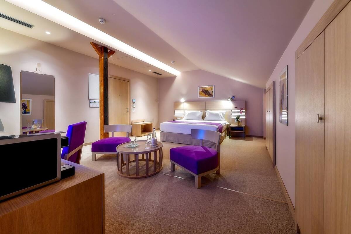 Ein Hotelzimmer mit einem Bett, zwei violetten Sesseln, einem kleinen runden Tisch, einem Fernseher und einem Kleiderschrank.
