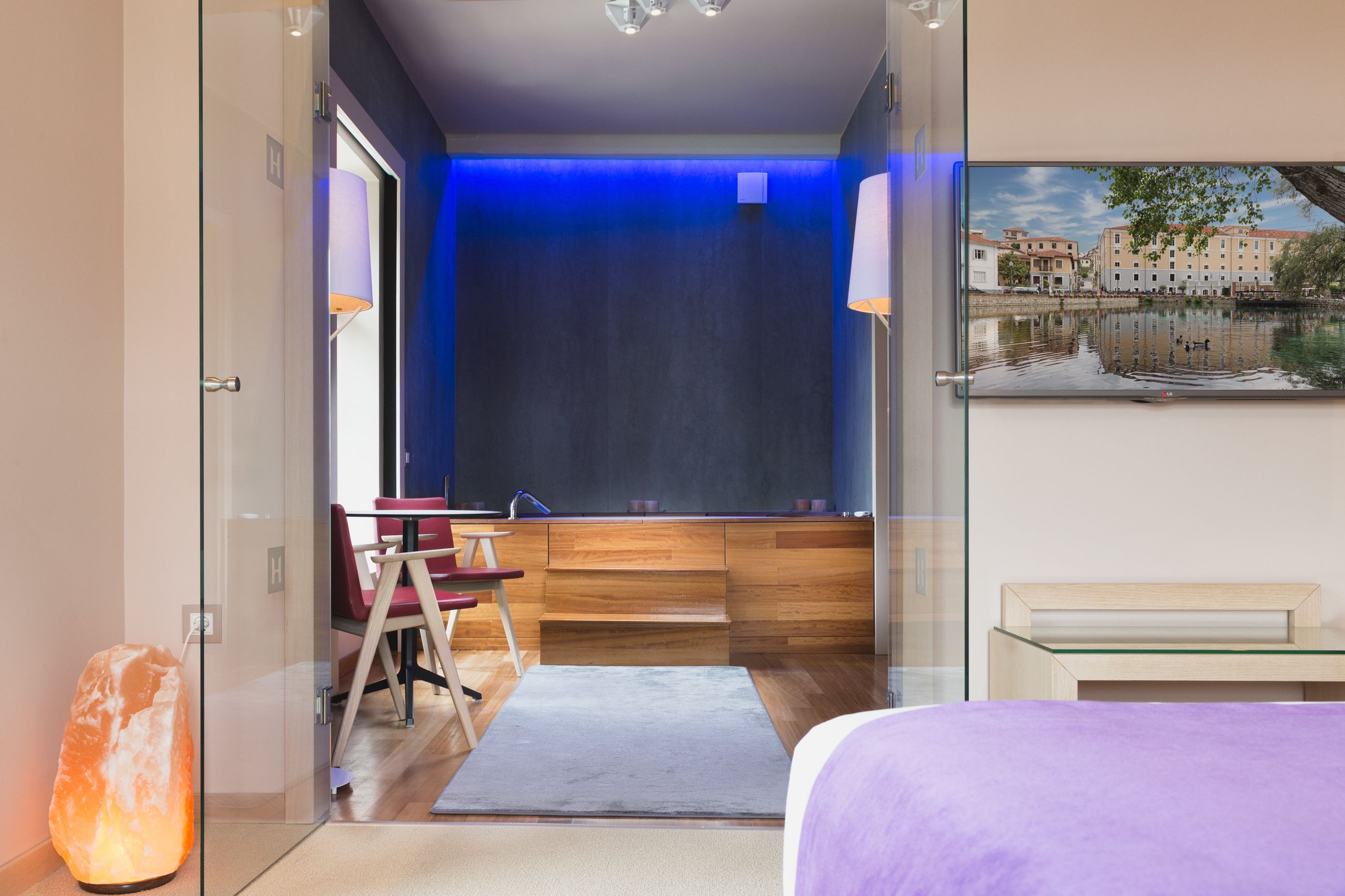 A modern hotel room with a glass door leading to a balcony, a bed with a purple bedspread, a wooden desk, and a chair.